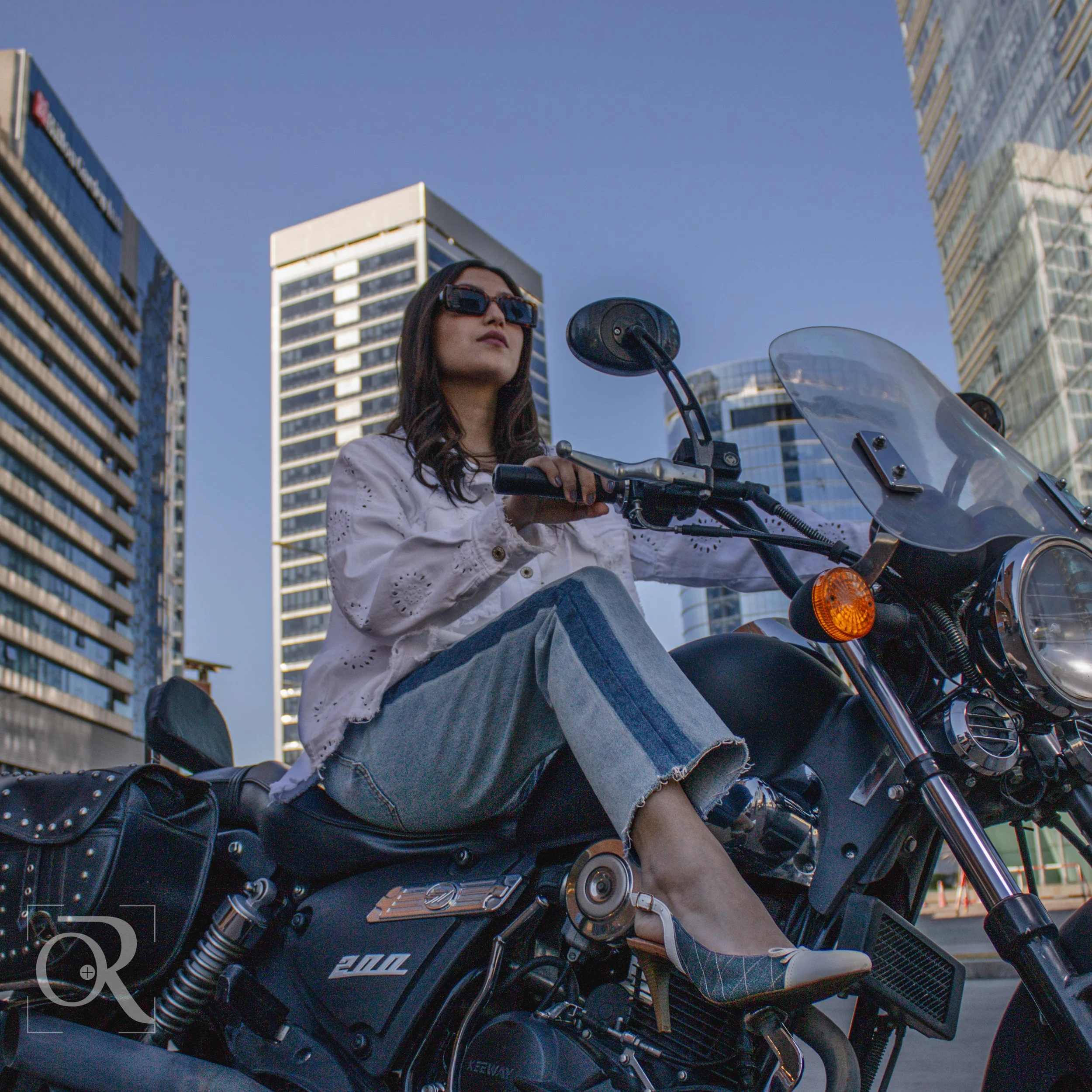 Mujer con gafas oscuras y blusa blanca sentada en una motocicleta negra en una ciudad con edificios altos de fondo