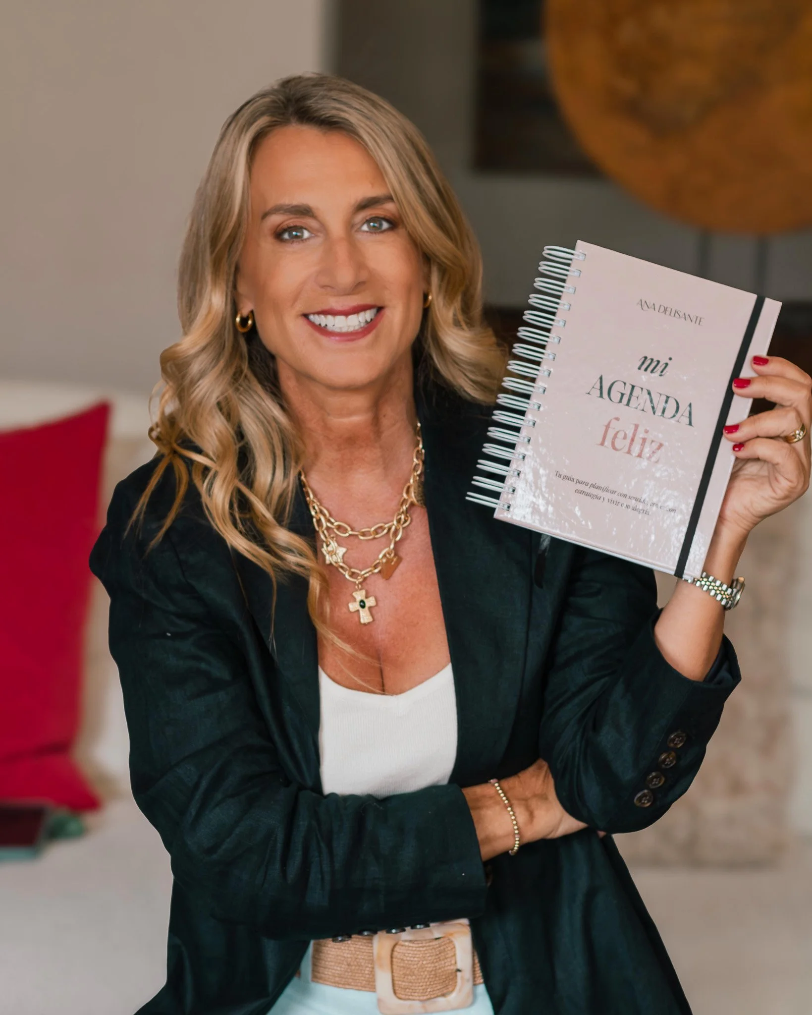 Mujer sonriendo, sosteniendo una agenda que dice 'mi agenda feliz' made in beige y negro, con fondo de hogar con cojines rojos.