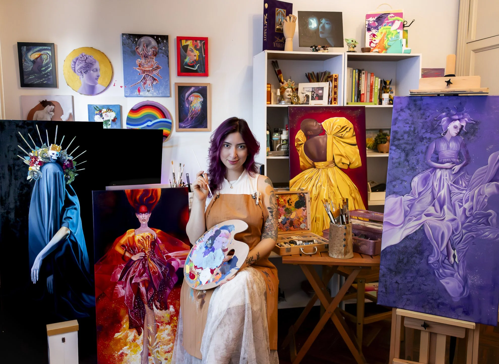 Artista en su estudio rodeada de pinturas de retratos y arte digital, sosteniendo una paleta y un pincel, con varias obras en proceso y en las paredes.