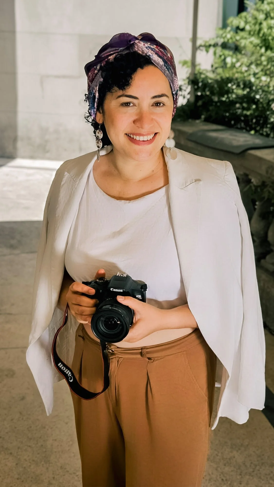 Mujer sonriente con cabello rizado, tocado de seda, blusa blanca, sudadera blanca y pantalones beige, sosteniendo una cámara Canon en un entorno al aire libre con plantas y paredes de piedra.
