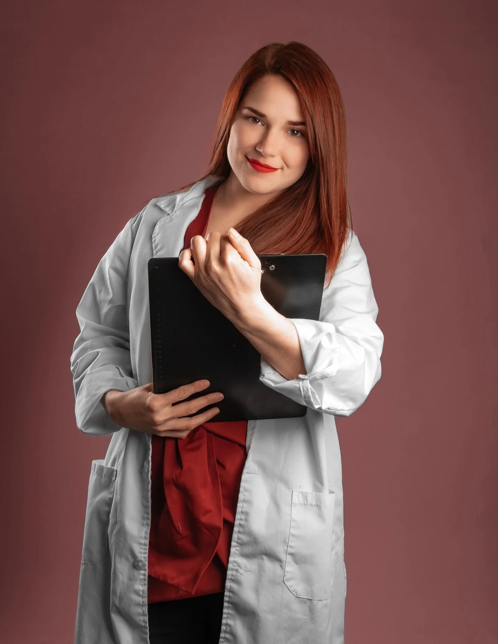 Mujer con cabello rojo, vestida con bata blanca, sosteniendo una carpeta negra, frente a un fondo marrón.