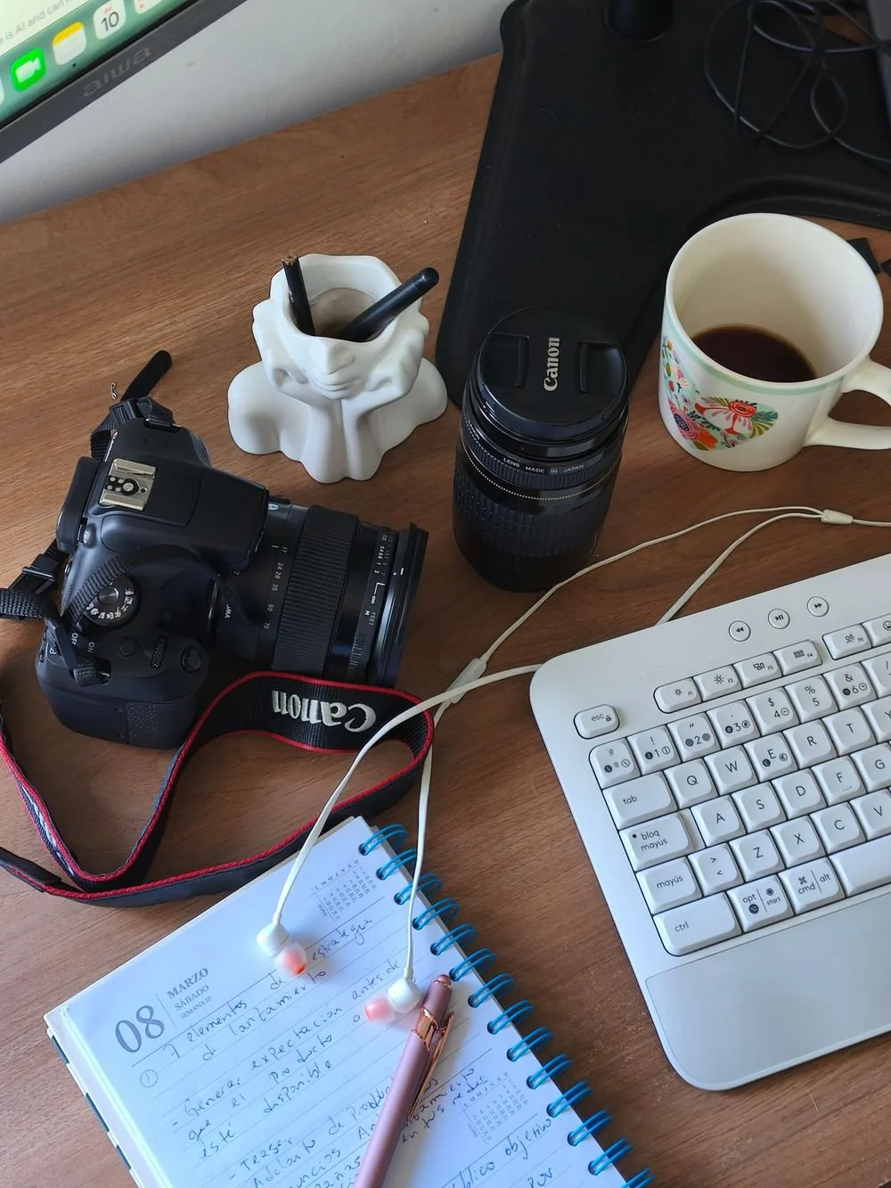 Mesa de trabajo con cámara Canon, lente, taza con café, computadora portátil, cuaderno de notas, bolígrafo y auriculares.