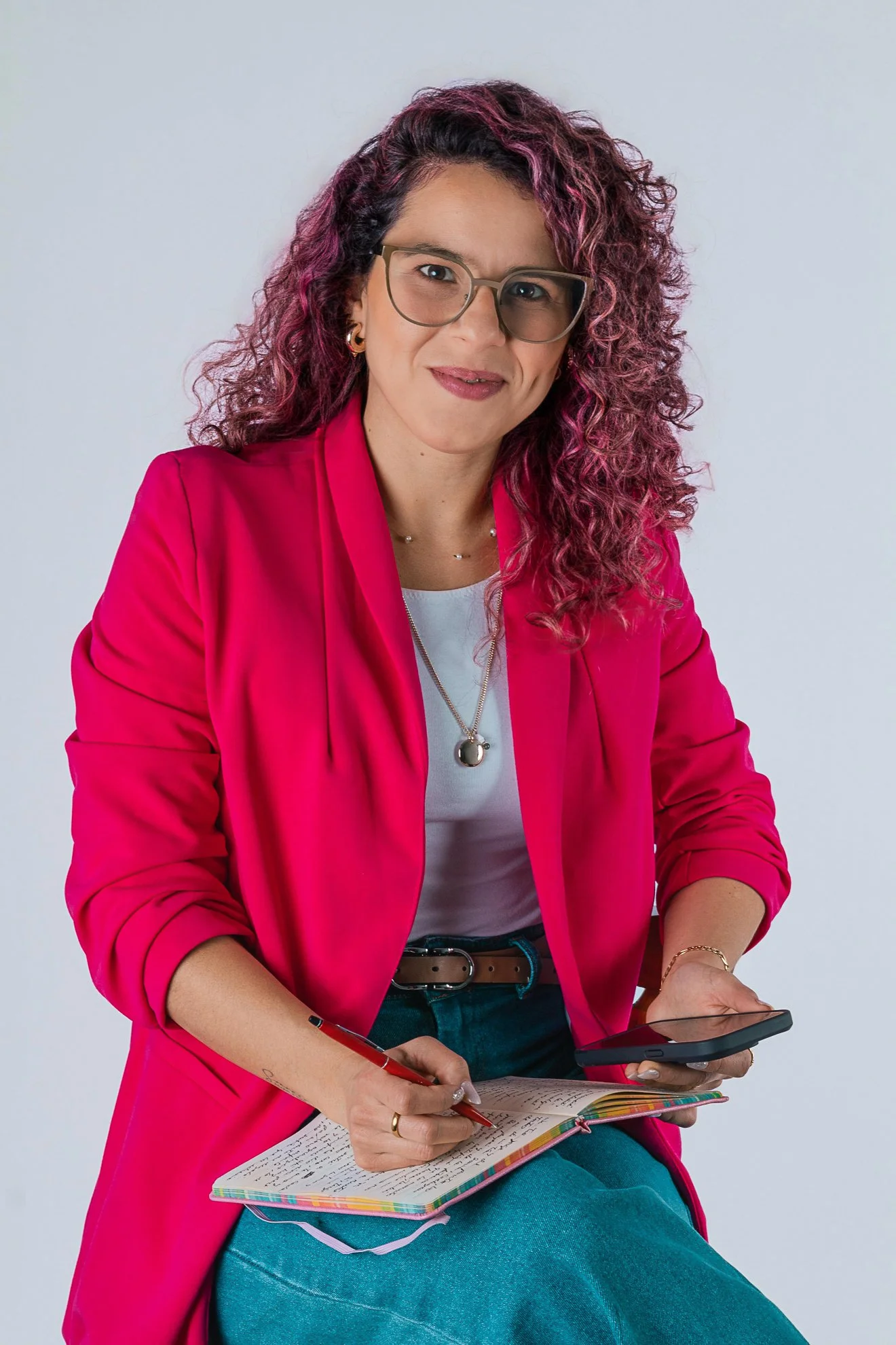 Mujer con cabello rizado y teñido de morado, con gafas, vestida con blusa blanca y chaqueta rosa, sosteniendo un teléfono móvil y una libreta con notas, sentada contra fondo gris.
