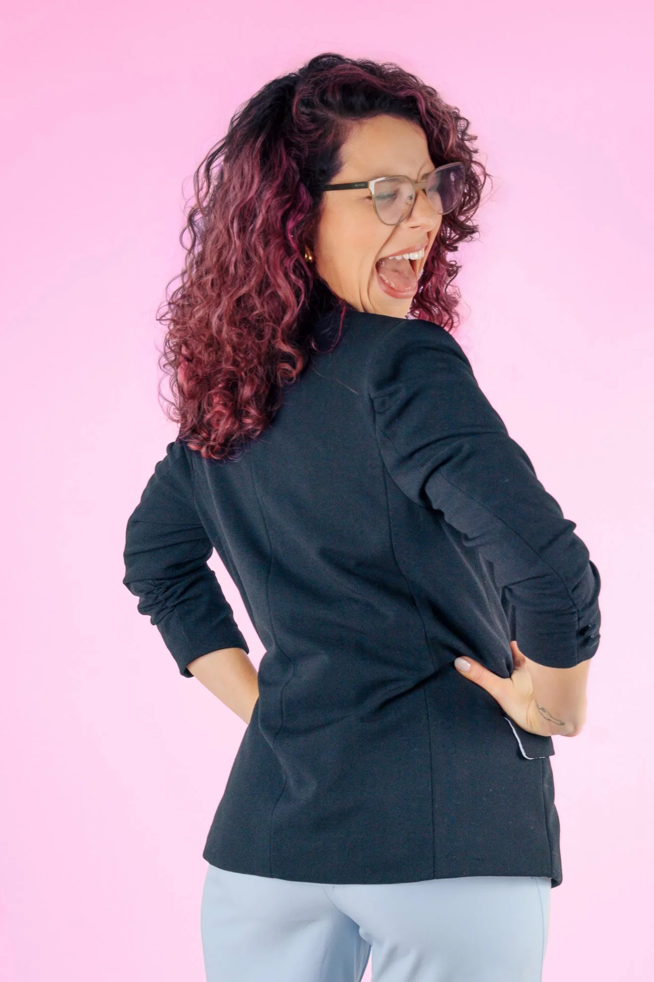Mujer con cabello rizado con mechones rosas, gafas, camisa negra, gesticulando y sonriendo con fondo rosa