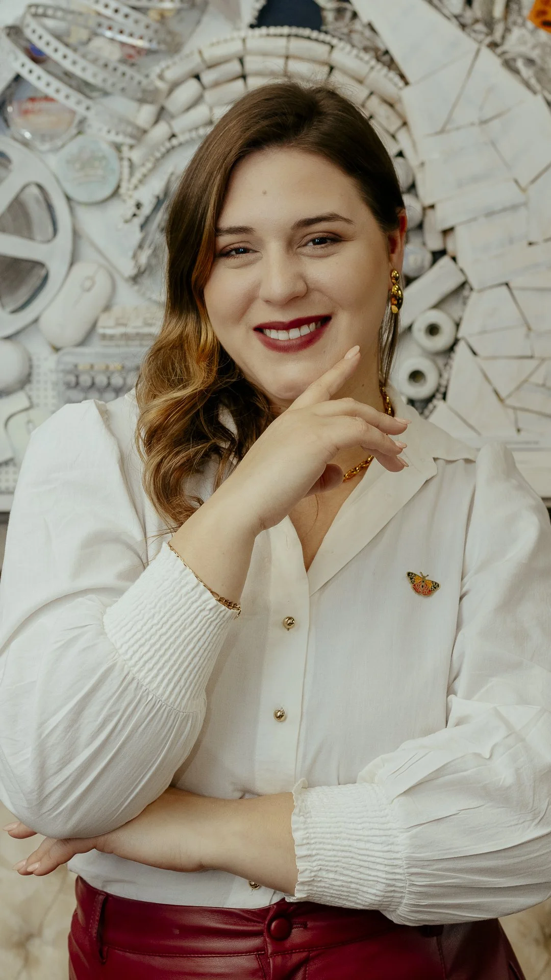 Retrato de una mujer sonriendo, usando blusa blanca y pendientes dorados, con fondo decorativo de objetos blancos y papel.