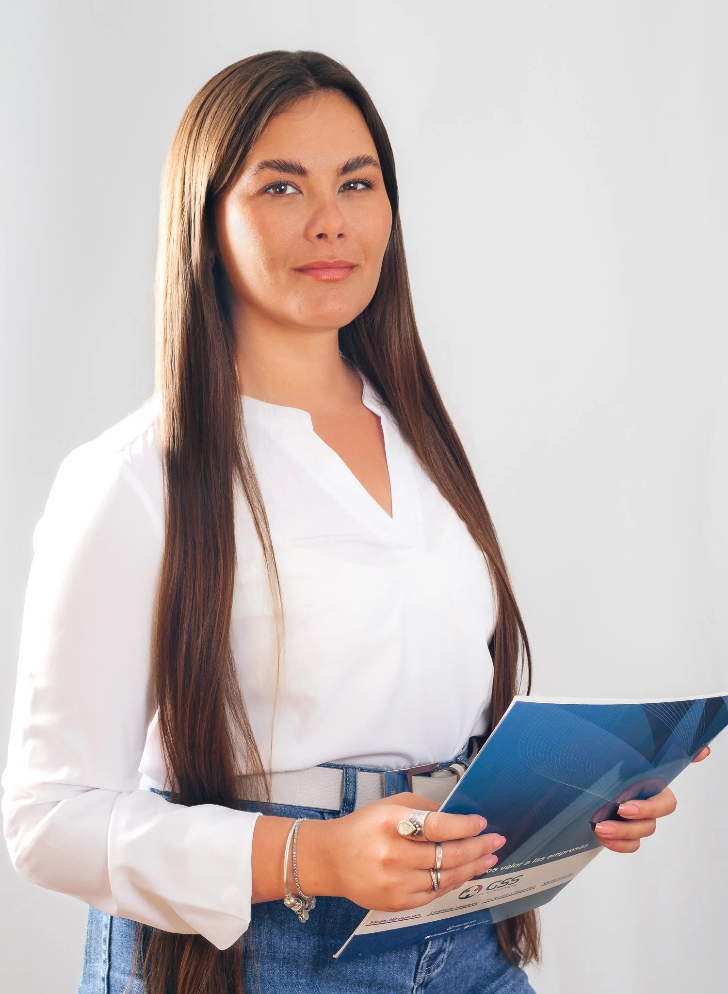 Mujer con cabello largo y camiseta blanca sosteniendo un folleto azul frente a fondo blanco.