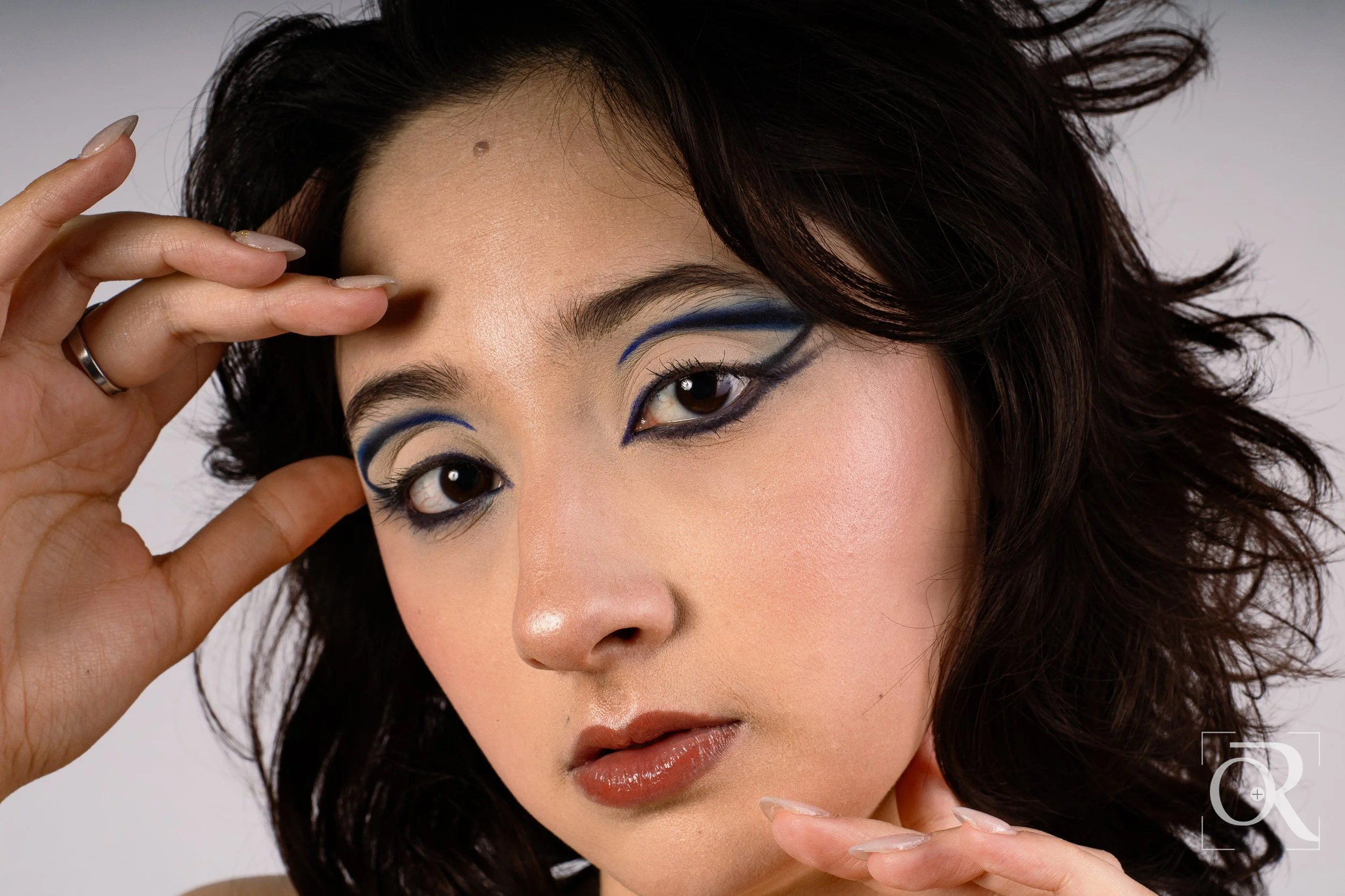 Retrato de una mujer con maquillaje artístico en los ojos, con cabello rizado oscuro y piel clara, posando con una expresión enérgica y manos cerca del rostro.