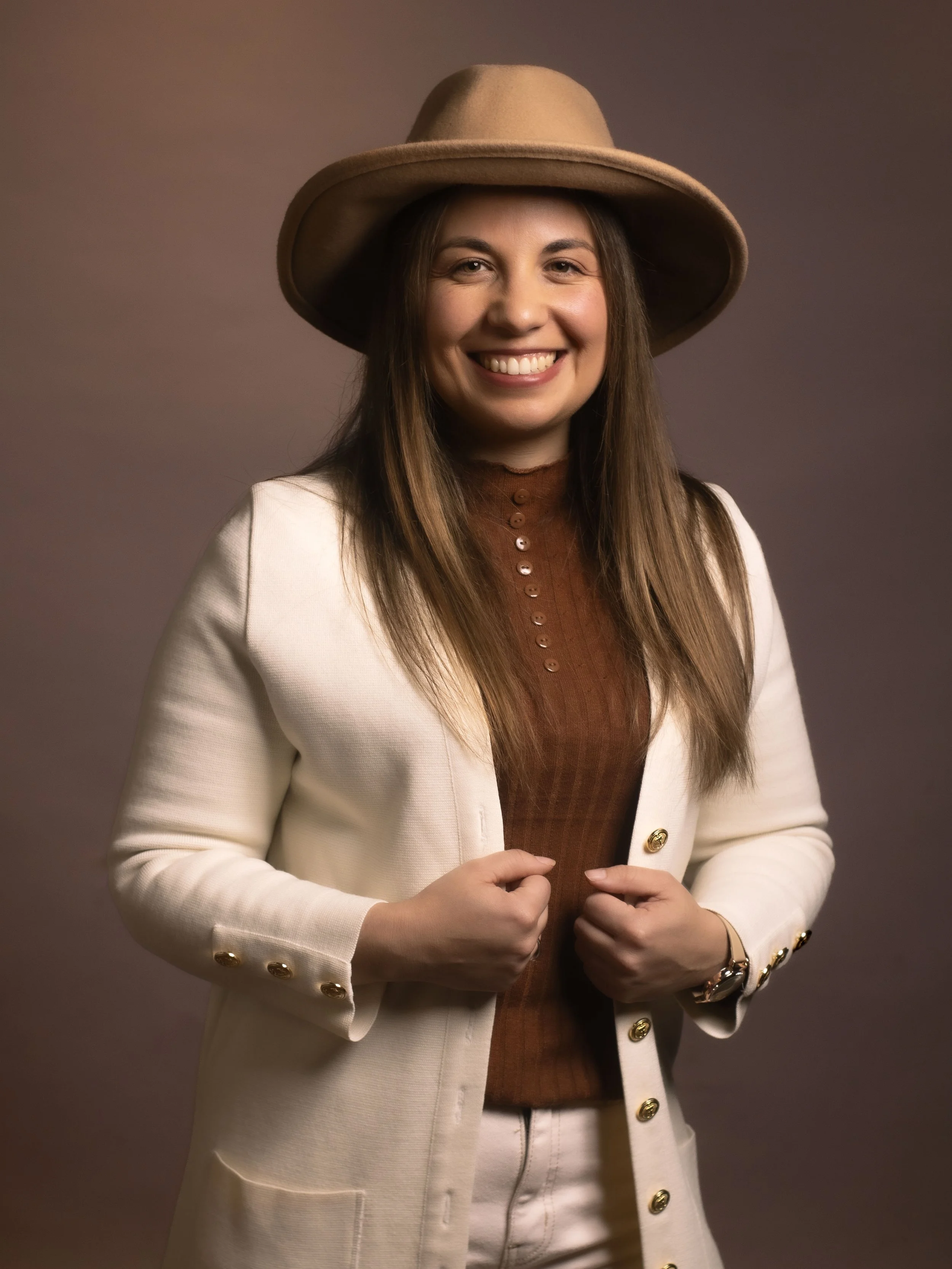 Una mujer sonriente con sombrero beige, chaqueta blanca y blusa marrón, posando con confianza.