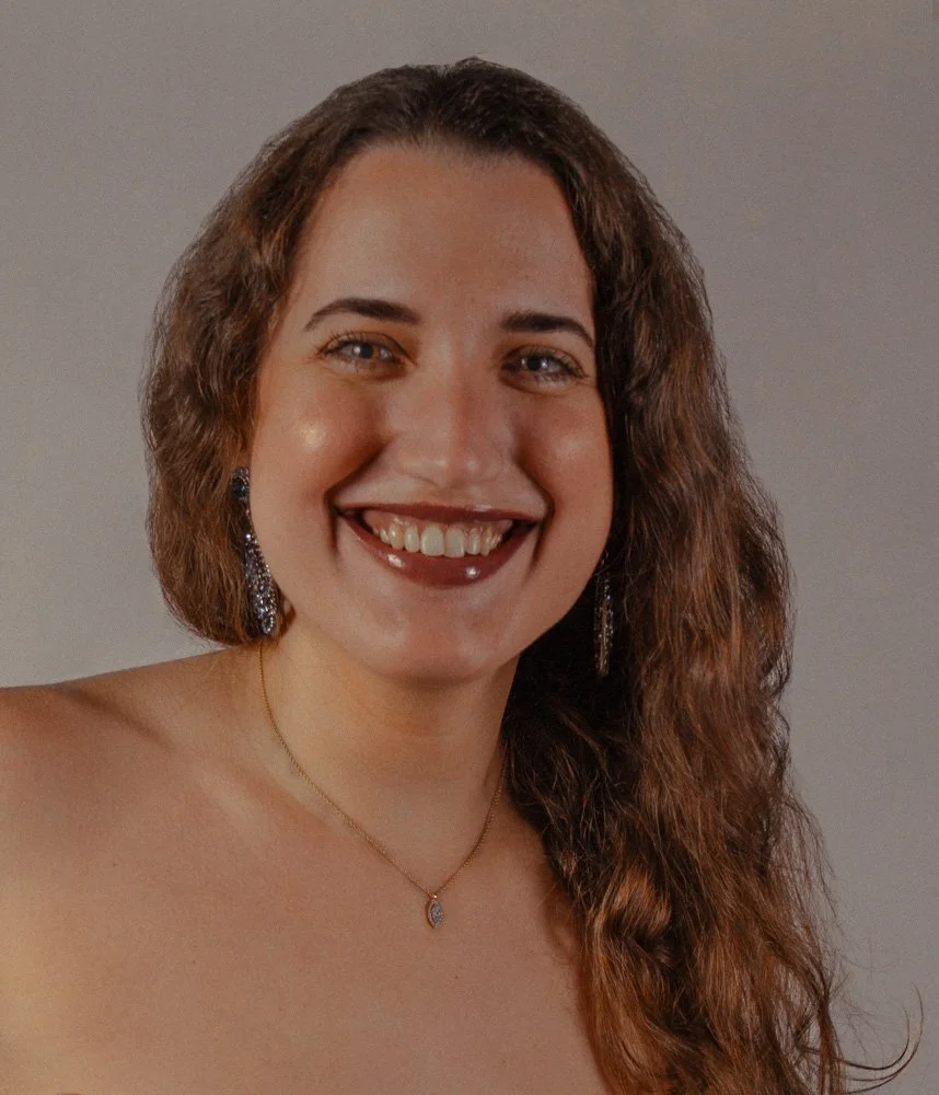 Retrato de una mujer sonriente con cabello rizado y largo, usando pendientes de aro grandes, collar dorado con colgante y maquillaje natural contra fondo gris.