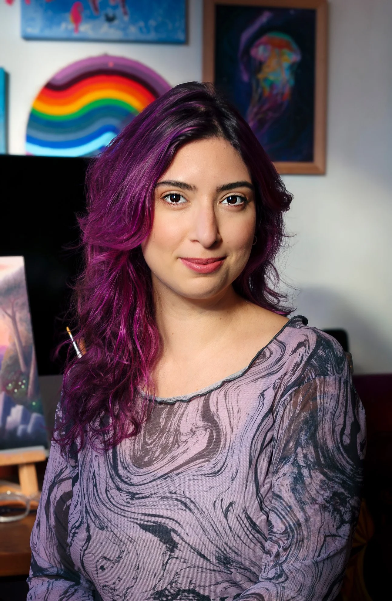 Retrato de una mujer con cabello largo y rizado teñido de tonos morados y rosados, con fondo de obras de arte coloridas.