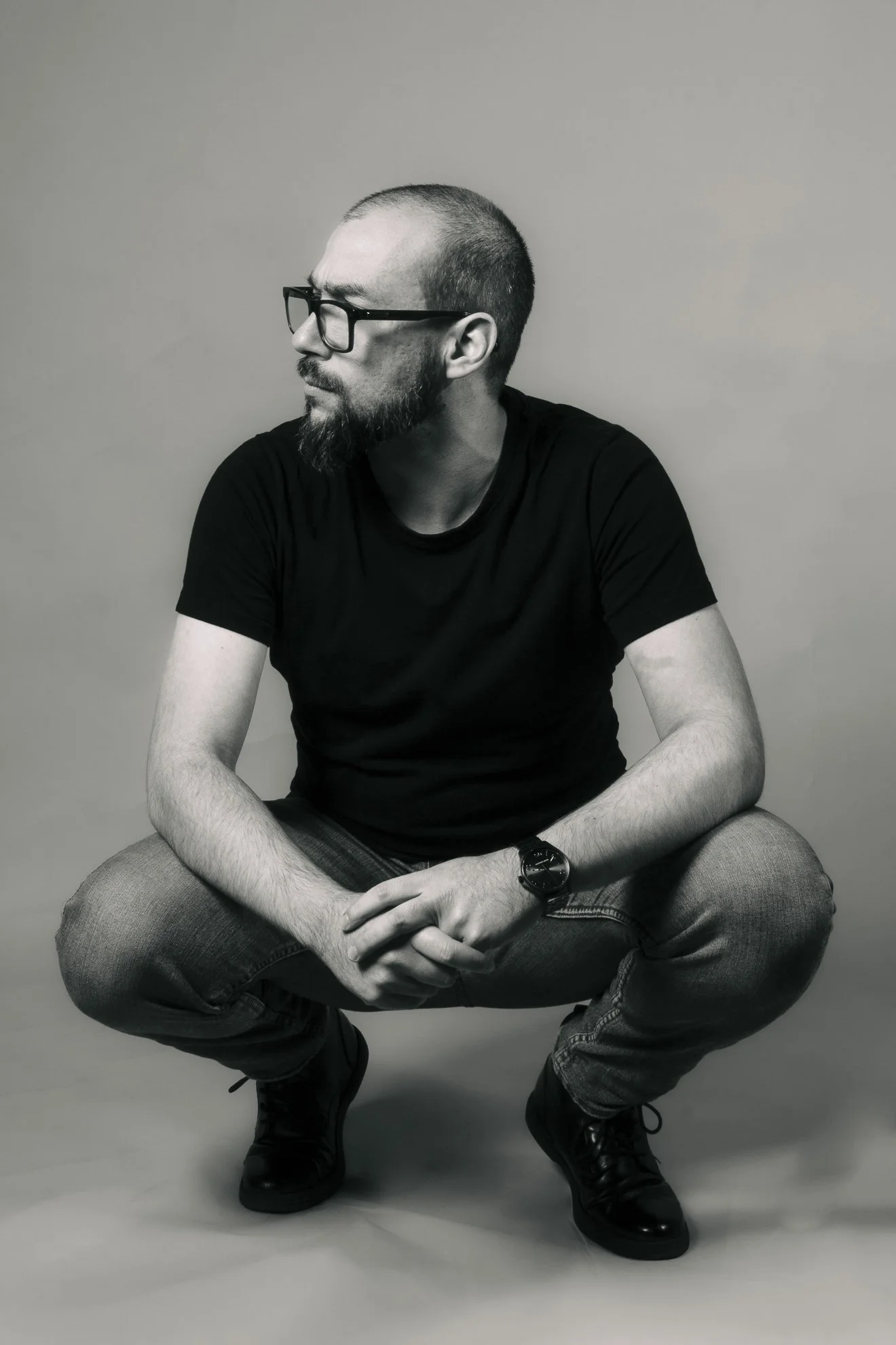 Hombre con barba, gafas y camiseta negra, sentado en cuclillas contra fondo gris, mirando hacia la izquierda.