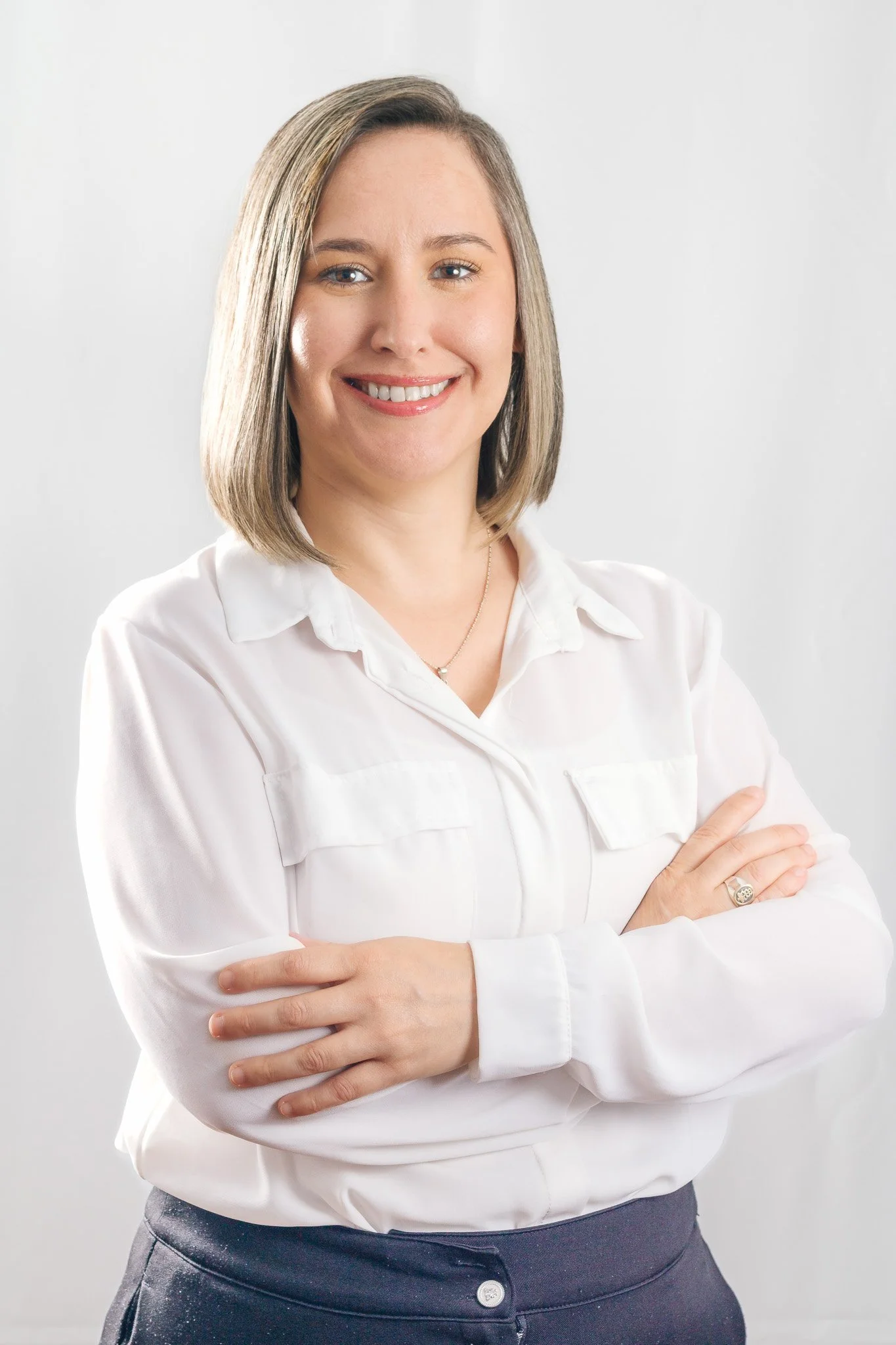 Mujer sonriendo con brazos cruzados, usando blusa blanca y pantalón oscuro, contra fondo blanco.
