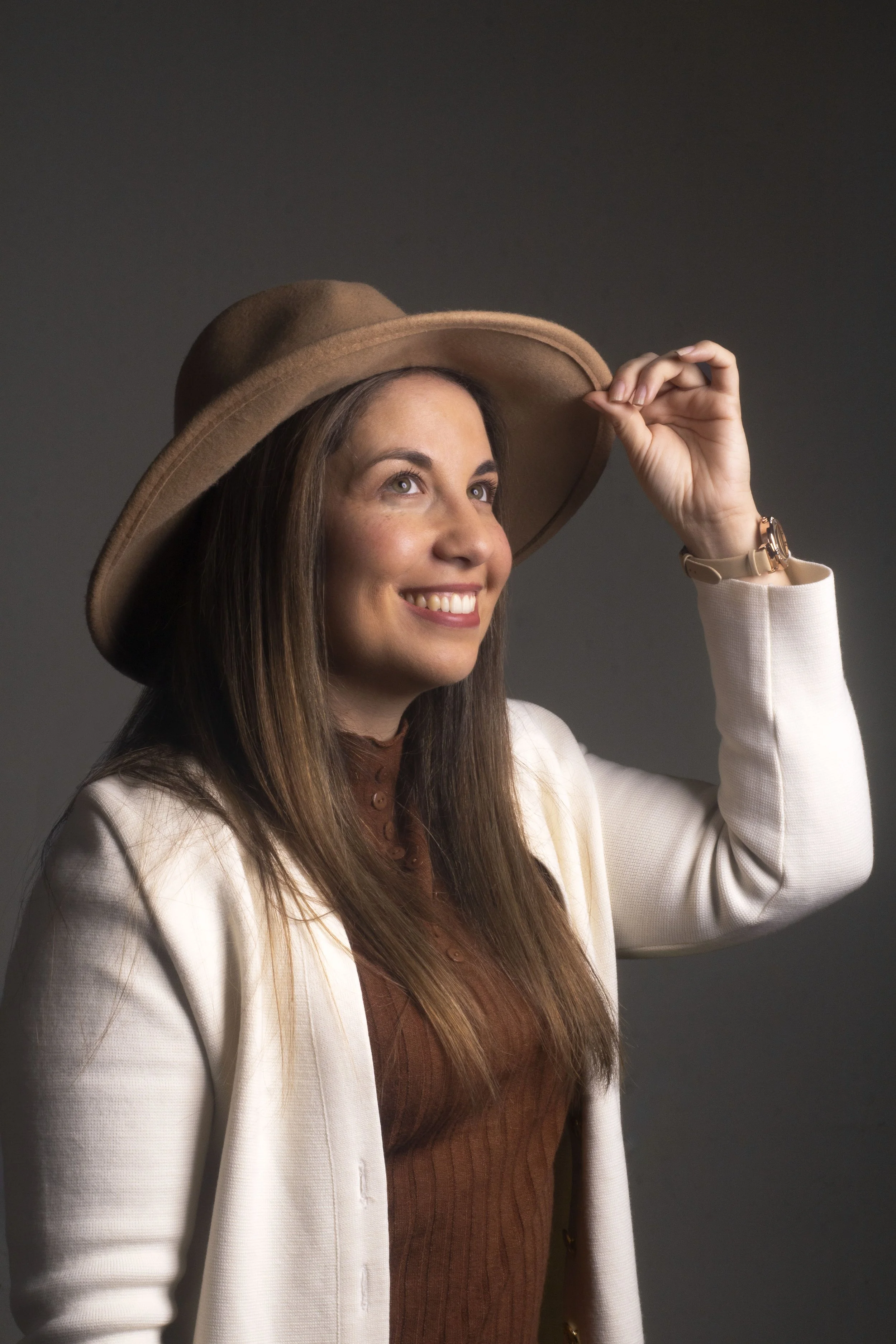 Mujer sonriendo, con sombrero y ropa formal, tocando su sombrero en un fondo gris oscuro.