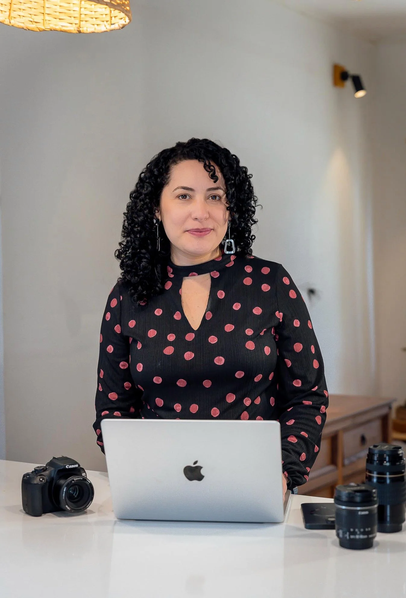Mujer con cabello rizado y oscuro, usando blusa negra con puntos rosados, de pie frente a una laptop en una mesa con una cámara y lentes fotográficos, en un ambiente interior.