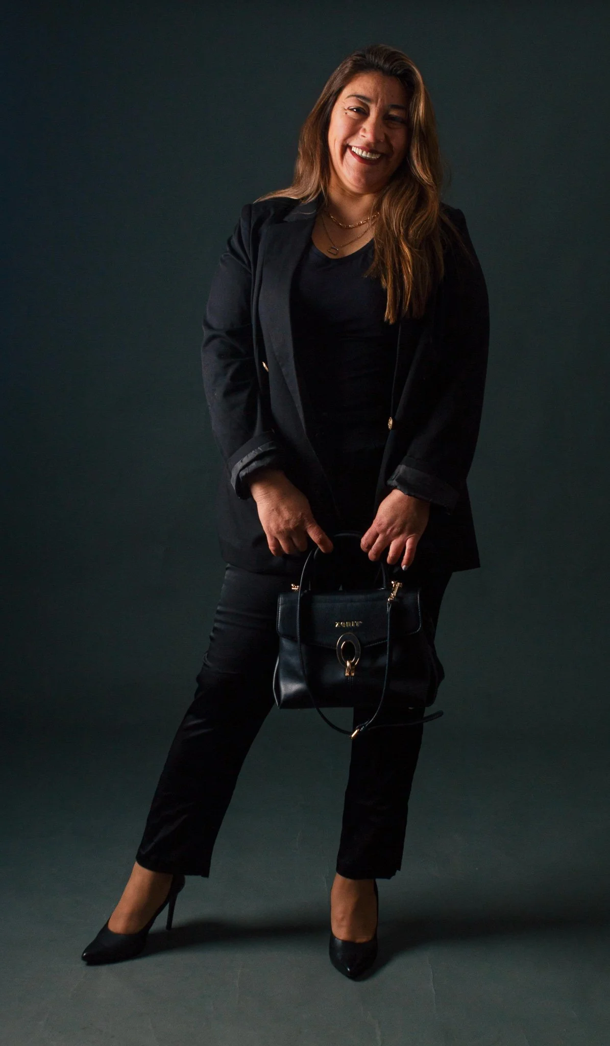 Mujer sonriendo, vestida con ropa negra, sosteniendo un bolso negro y usando tacones negros, contra un fondo oscuro.
