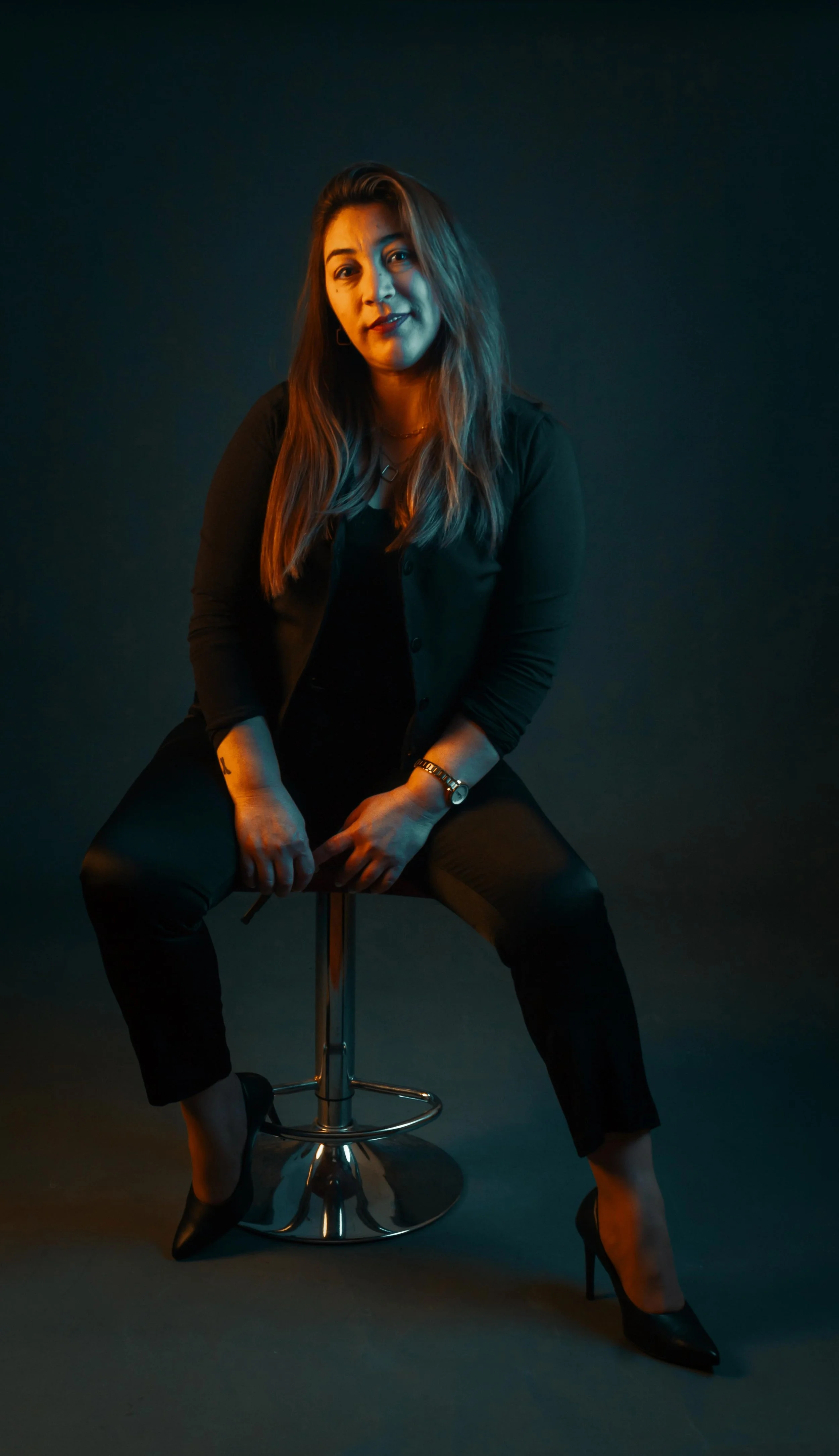 Mujer sentado en un taburete con fondo oscuro, vistiendo ropa negra y tacones, con iluminación de colores cálidos y fríos.