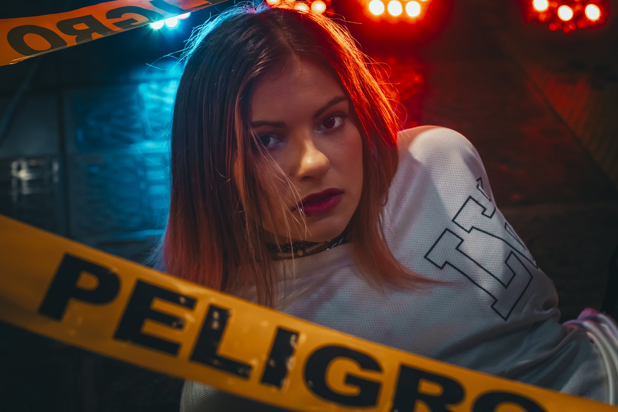 Joven mujer con cabello castaño y rubio, con expresión de susto o angustia, rodeada de cinta de aviso policial en un ambiente oscuro y con luces de neón de colores.