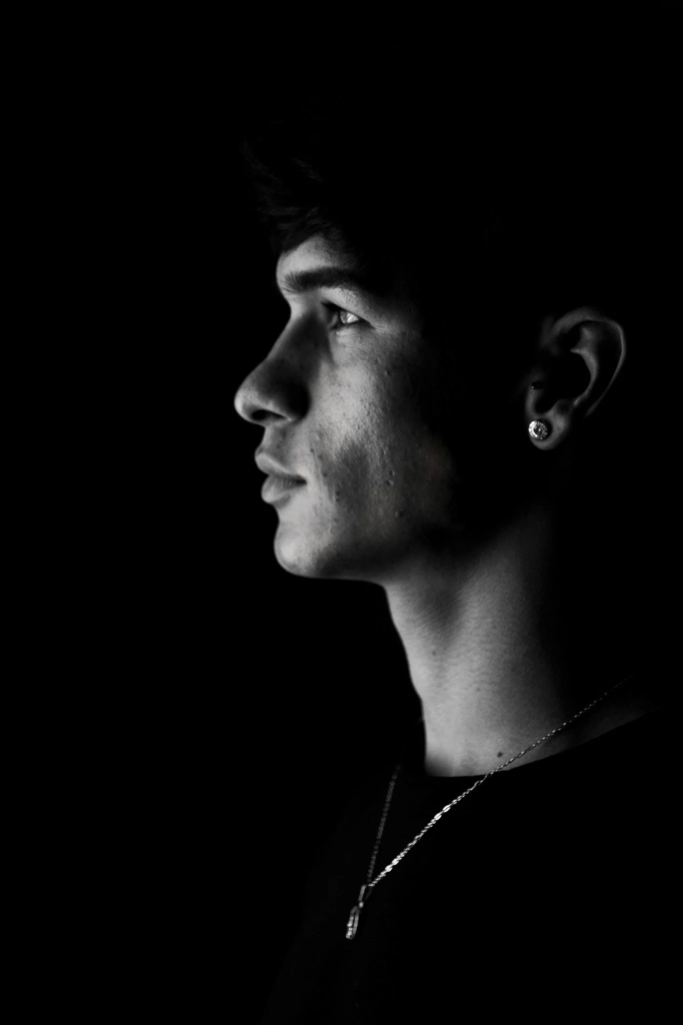 Retrato en blanco y negro de un joven mirando hacia la izquierda, con cabello oscuro, pendiente en la oreja y collar. Fondo negro.
