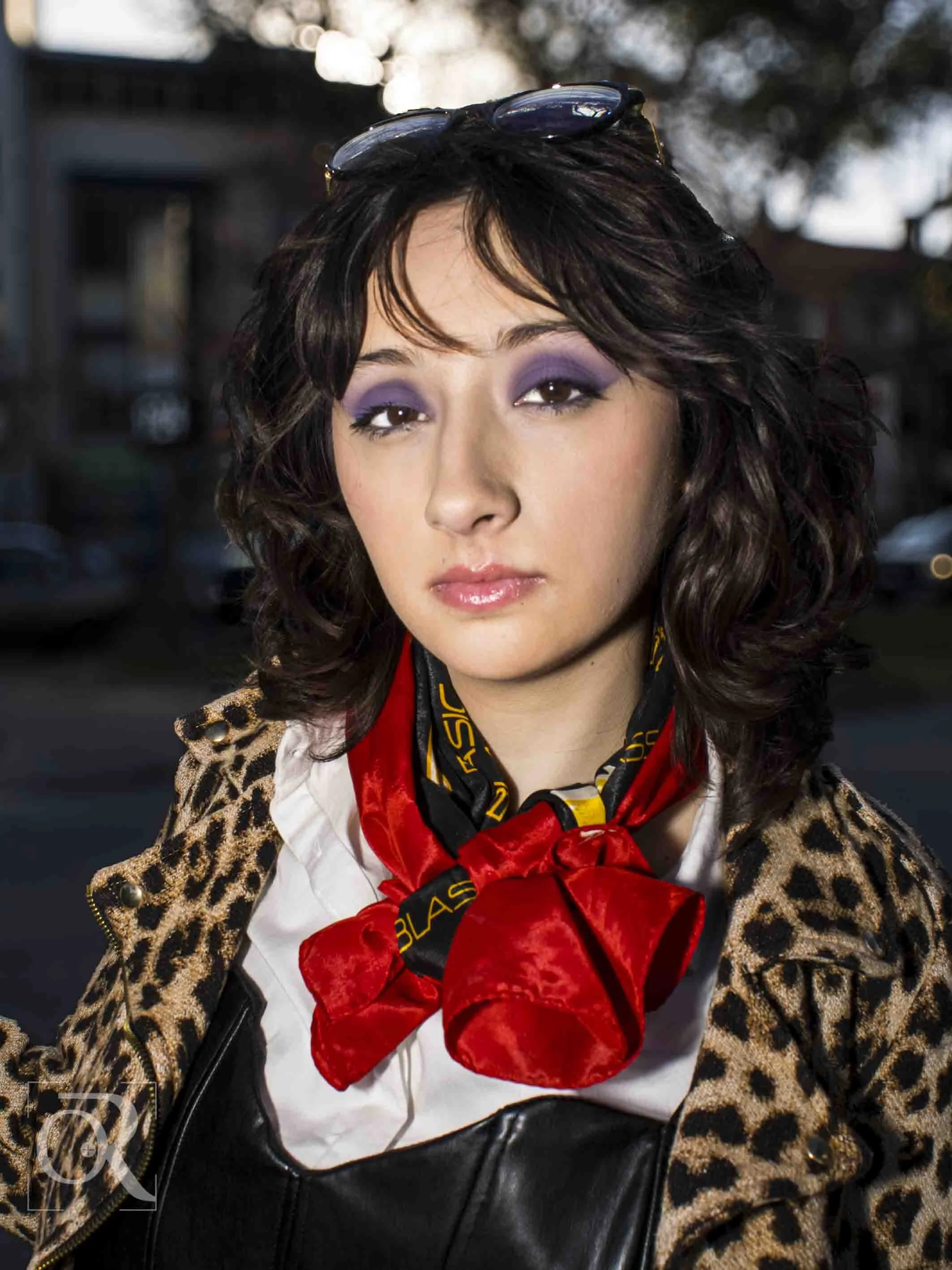 Retrato de una mujer con cabello rizado, lentes en la cabeza, maquillaje morado y labios rosados, con chaqueta de leopardo y pañuelo rojo