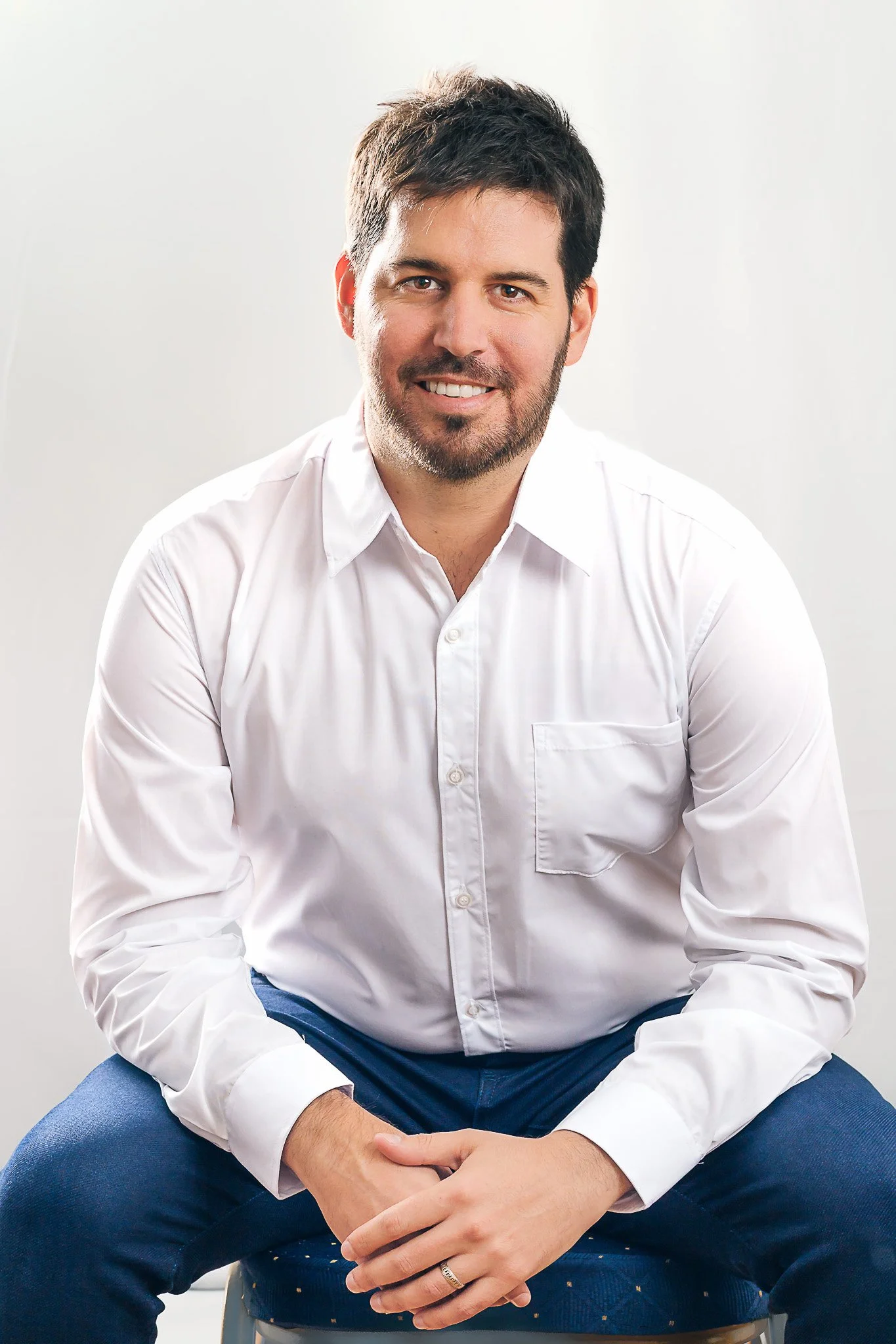 Un hombre con barba, cabello oscuro y camisa blanca, sentado en una silla, sonriendo frente a un fondo blanco.