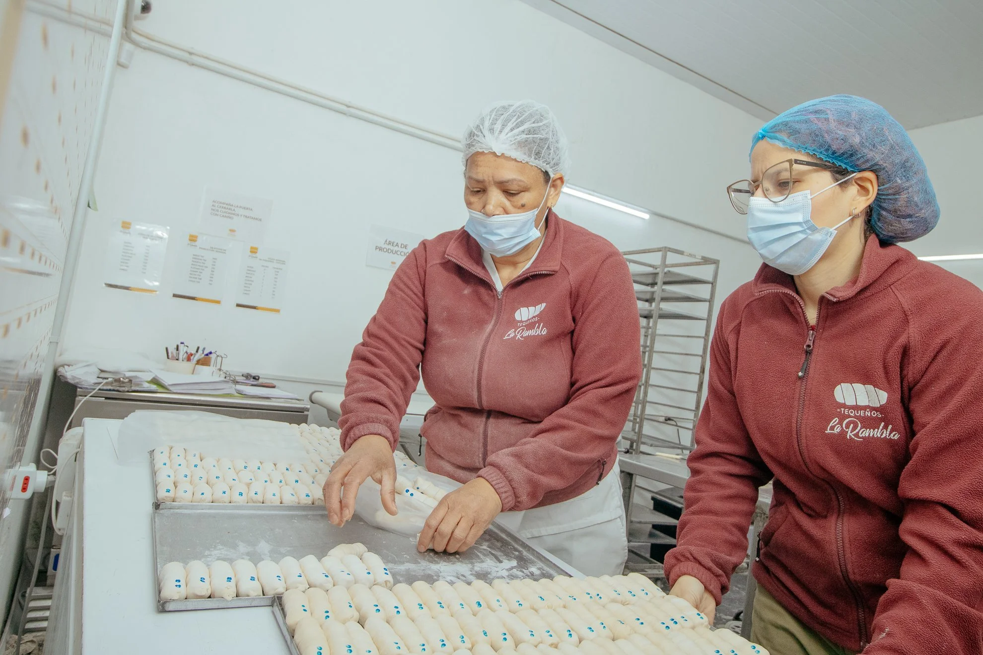 Dos mujeres trabajando en una panadería, revisando masa de pan con gorros y mascarillas, con estantes de productos en el fondo.