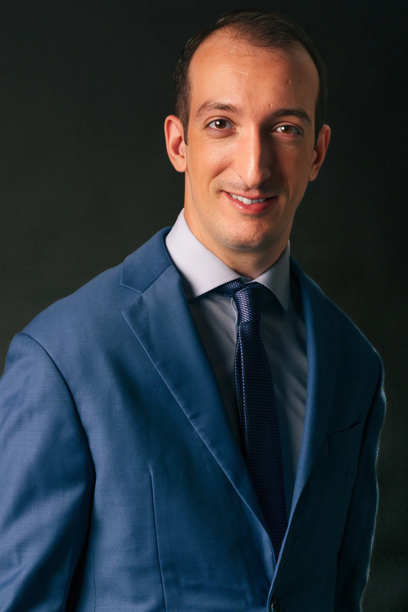 Hombre usando un traje azul con corbata, sonriendo y posando en un fondo oscuro.
