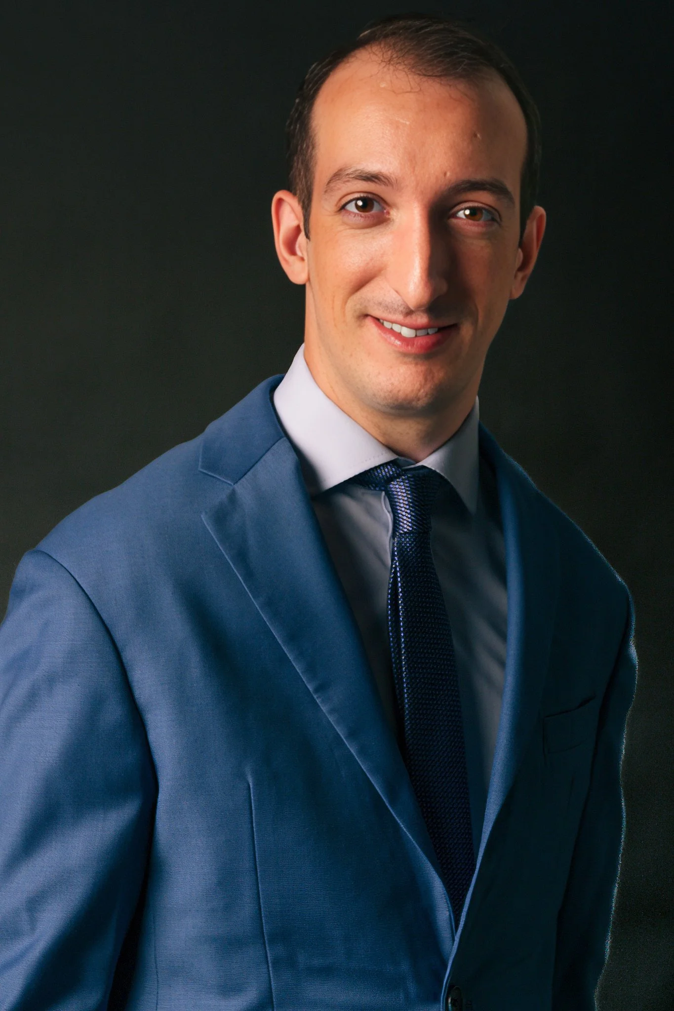 Retrato de un hombre sonriendo, lleva un saco azul, camisa blanca y corbata azul, fondo oscuro.