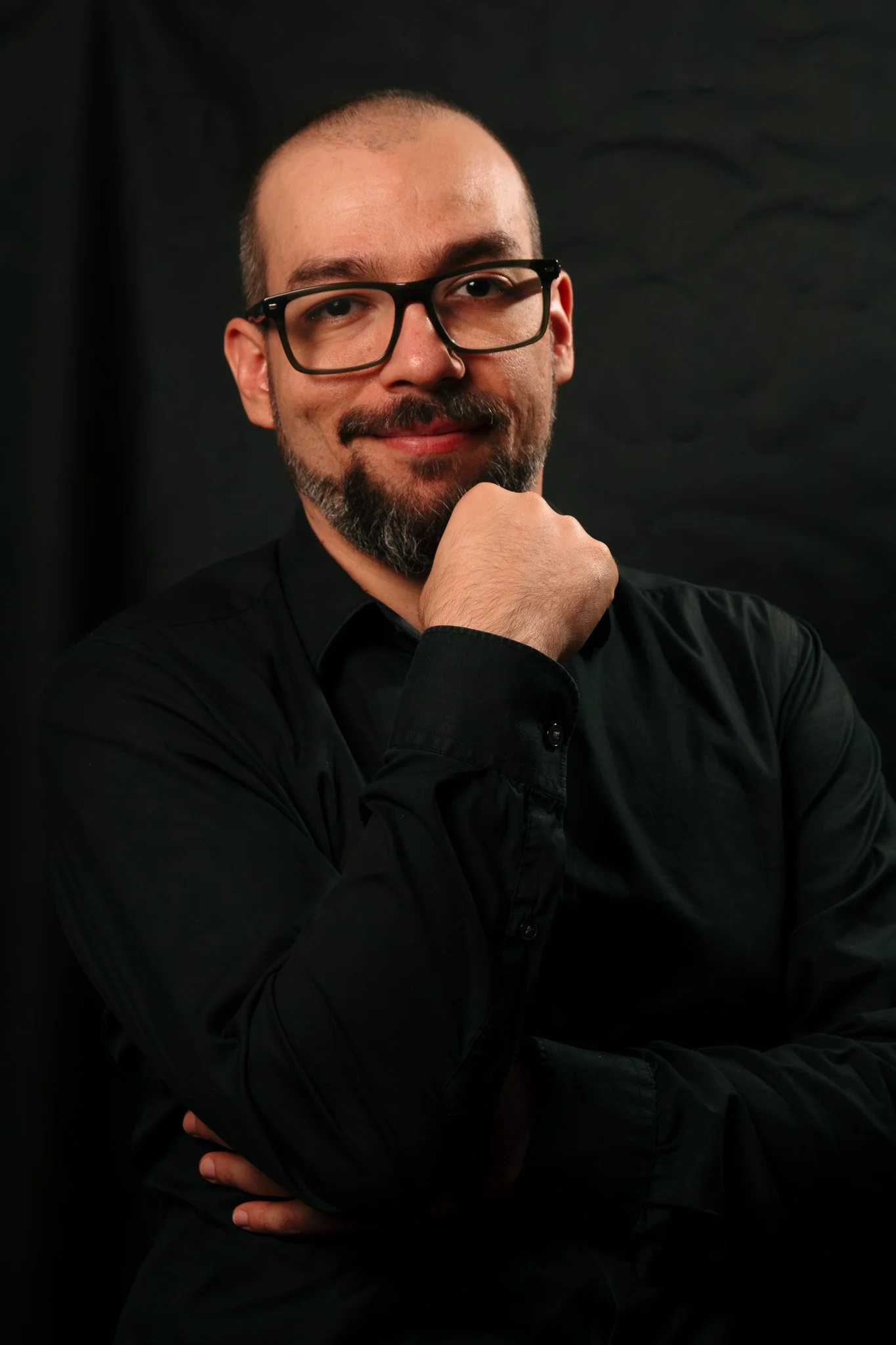 Hombre con barba, gafas negras y camisa negra, posando con la mano en la barbilla, sonriendo suavemente y mirando hacia la cámara, con fondo negro