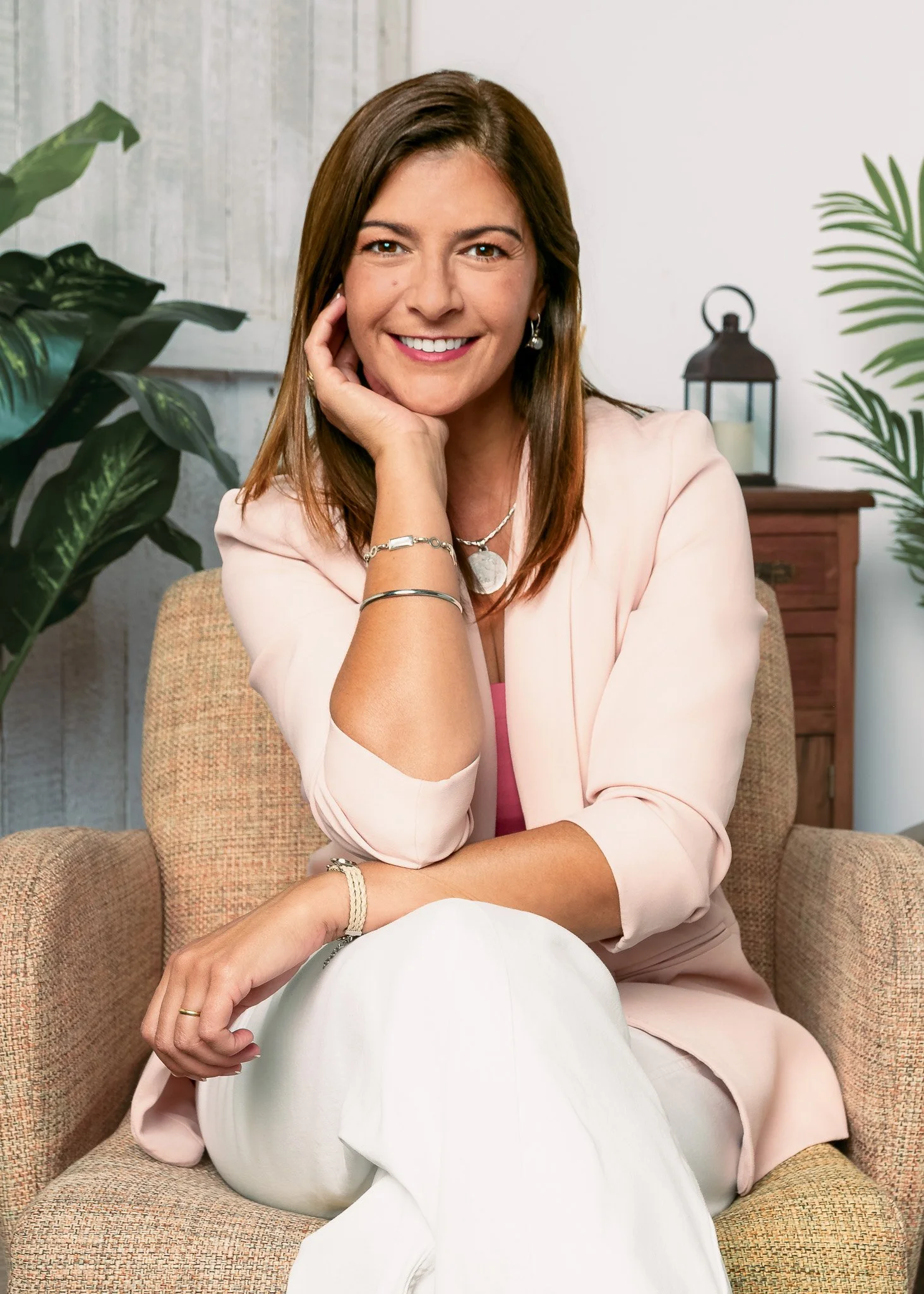 Retrato de una mujer con cabello castaño, sonriendo, usando blusa rosada y accesorios, sentada en un sillón beige en un entorno con plantas decorativas y muebles de madera.