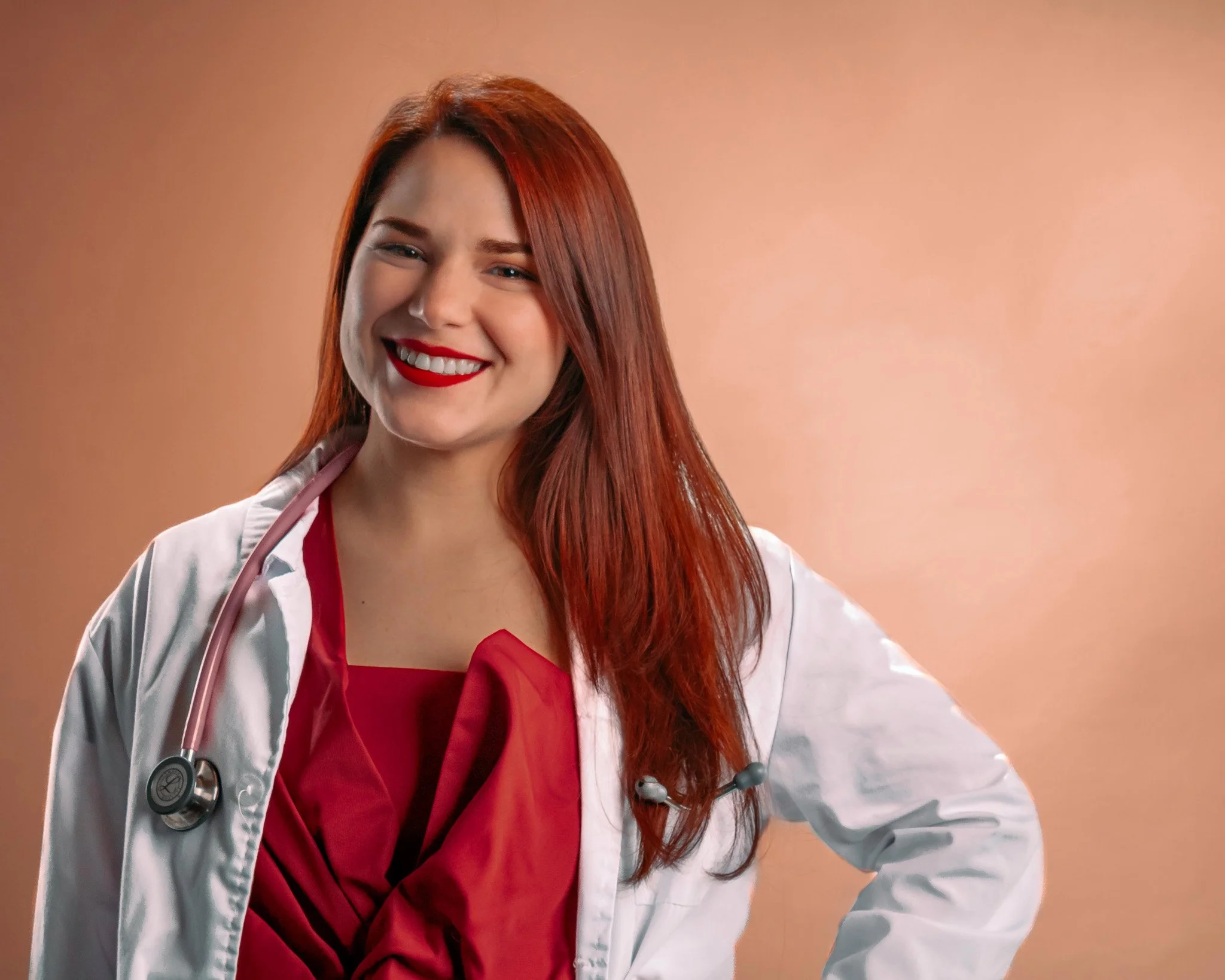 Una mujer con cabello rojo, sonriendo, usando bata blanca y un estetoscopio alrededor del cuello sobre fondo beige.