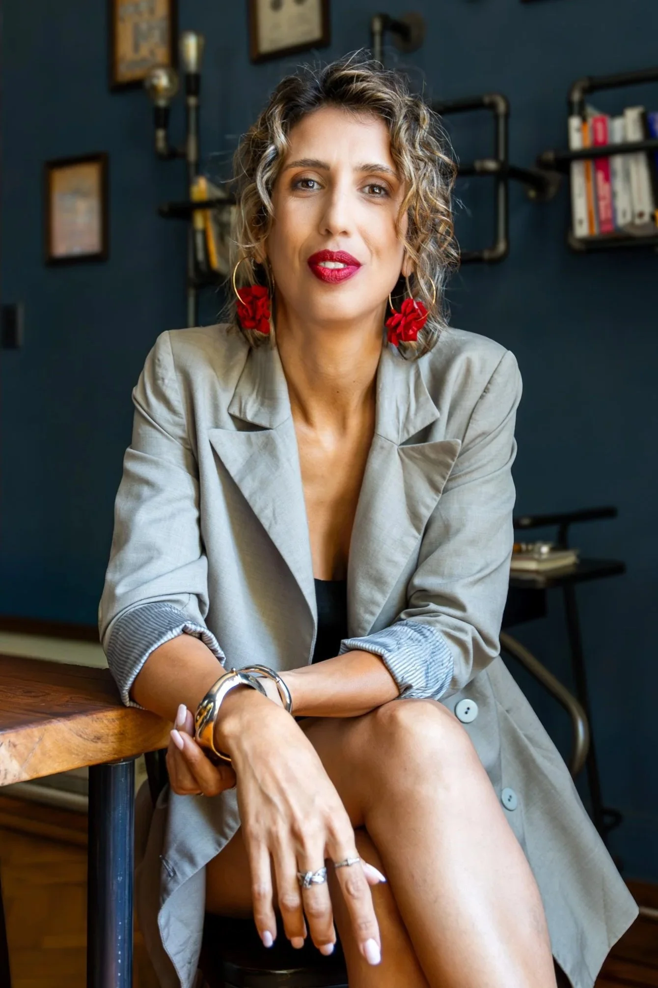 Mujer con cabello rizado y maquillaje rojo, usando un vestido negro y un saco gris, sentada en una mesa de madera en un ambiente moderno con paredes azules y estantes con libros
