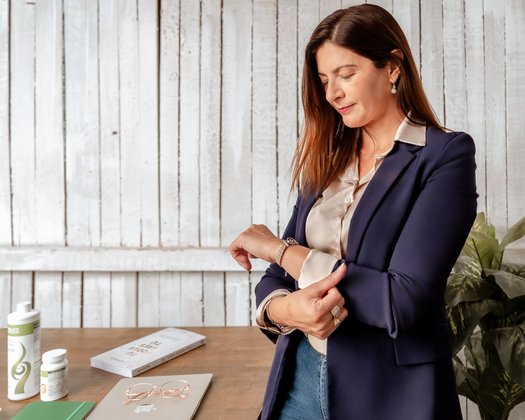 Mujer revisando su reloj en una mesa de madera, con gafas, libros, un bote de vitaminas y un suplemento, y una planta de interior en el fondo.