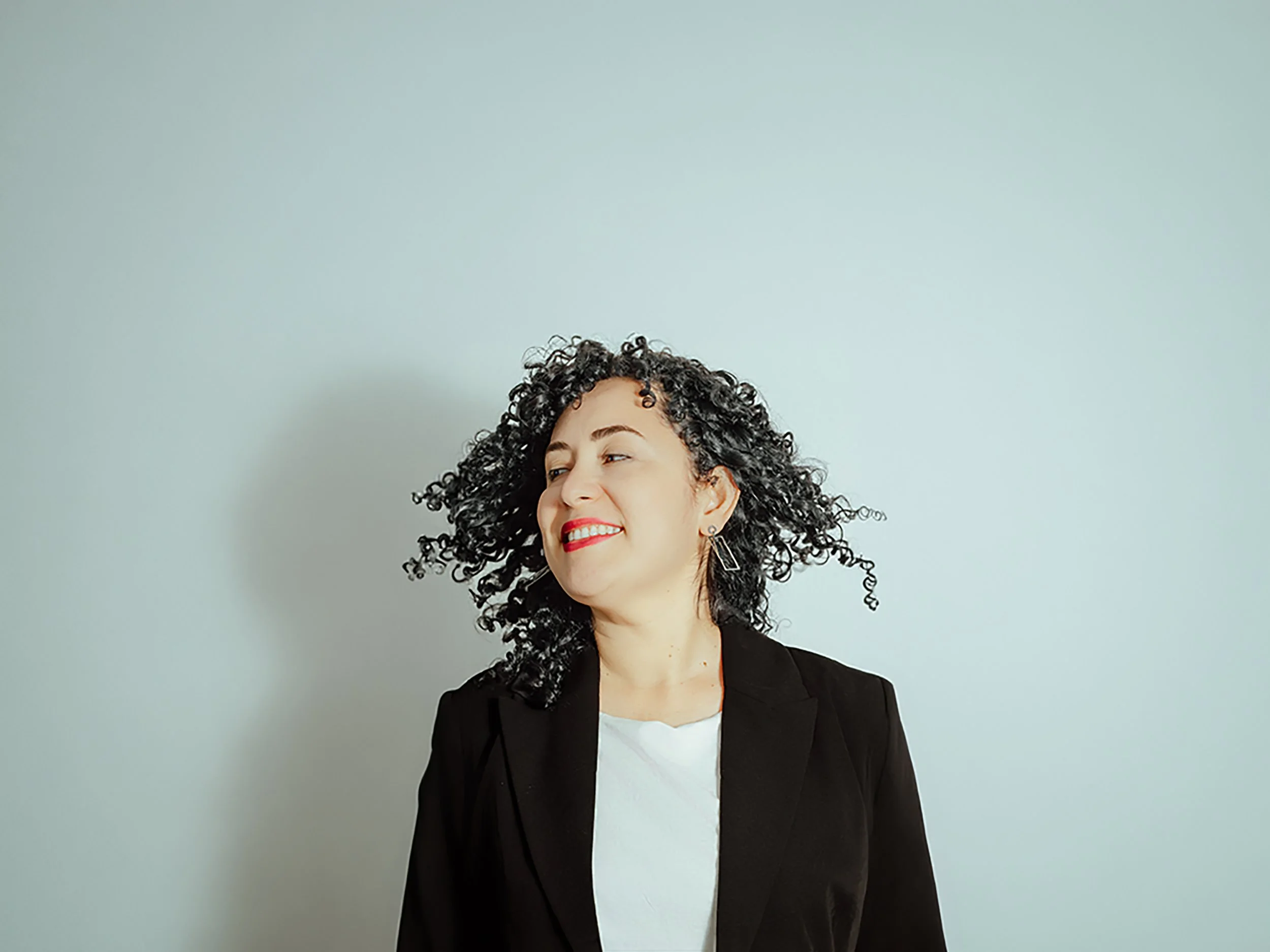 Mujer con cabello rizado, sonriendo, con blusa blanca y saco negro, de fondo gris claro.