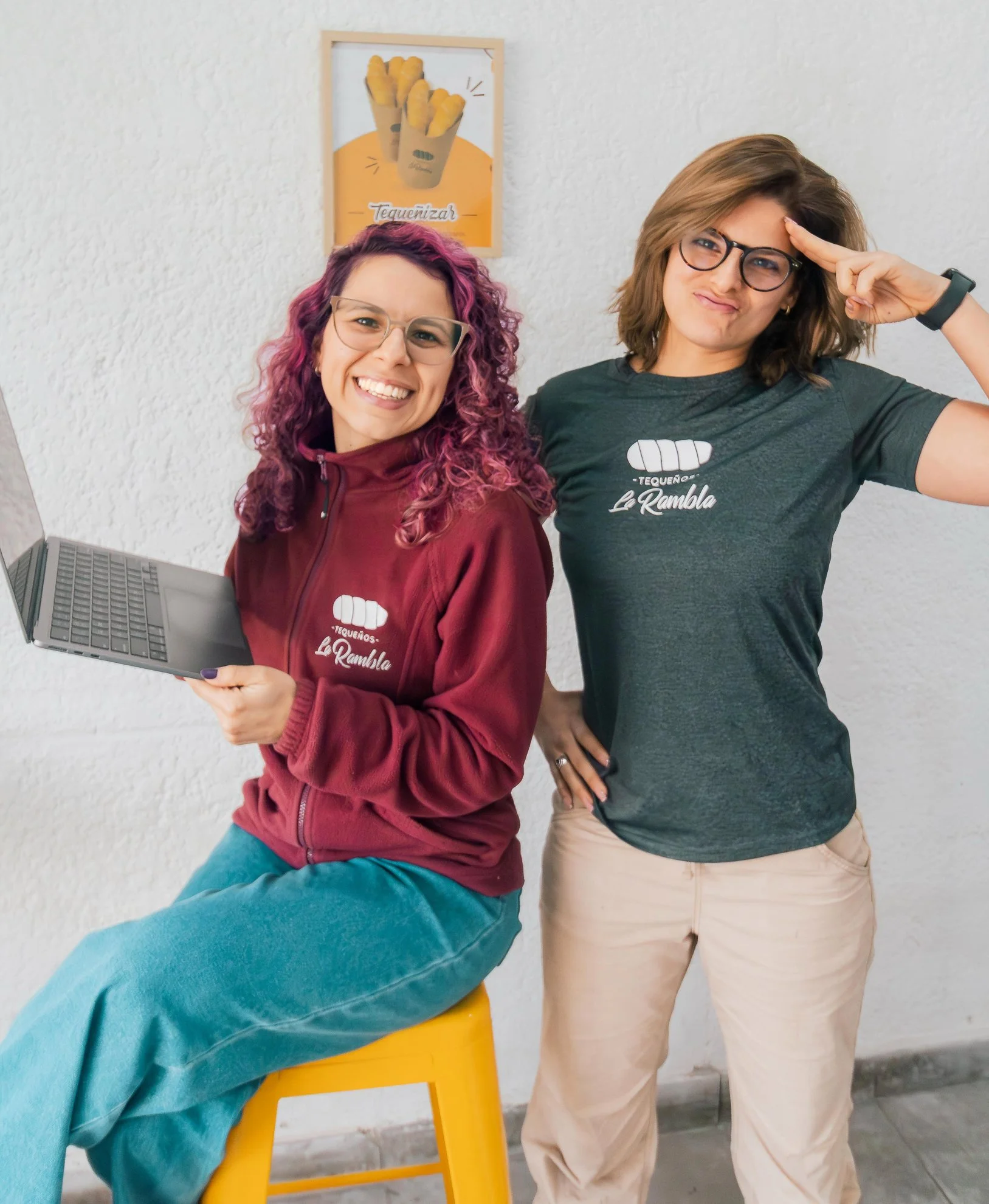 Dos mujeres jóvenes con gafas posando junto a una pared blanca, una de ellas sosteniendo una computadora portátil y ambas vestidas con camisetas que dicen 'Tequearnos La Rambla'.