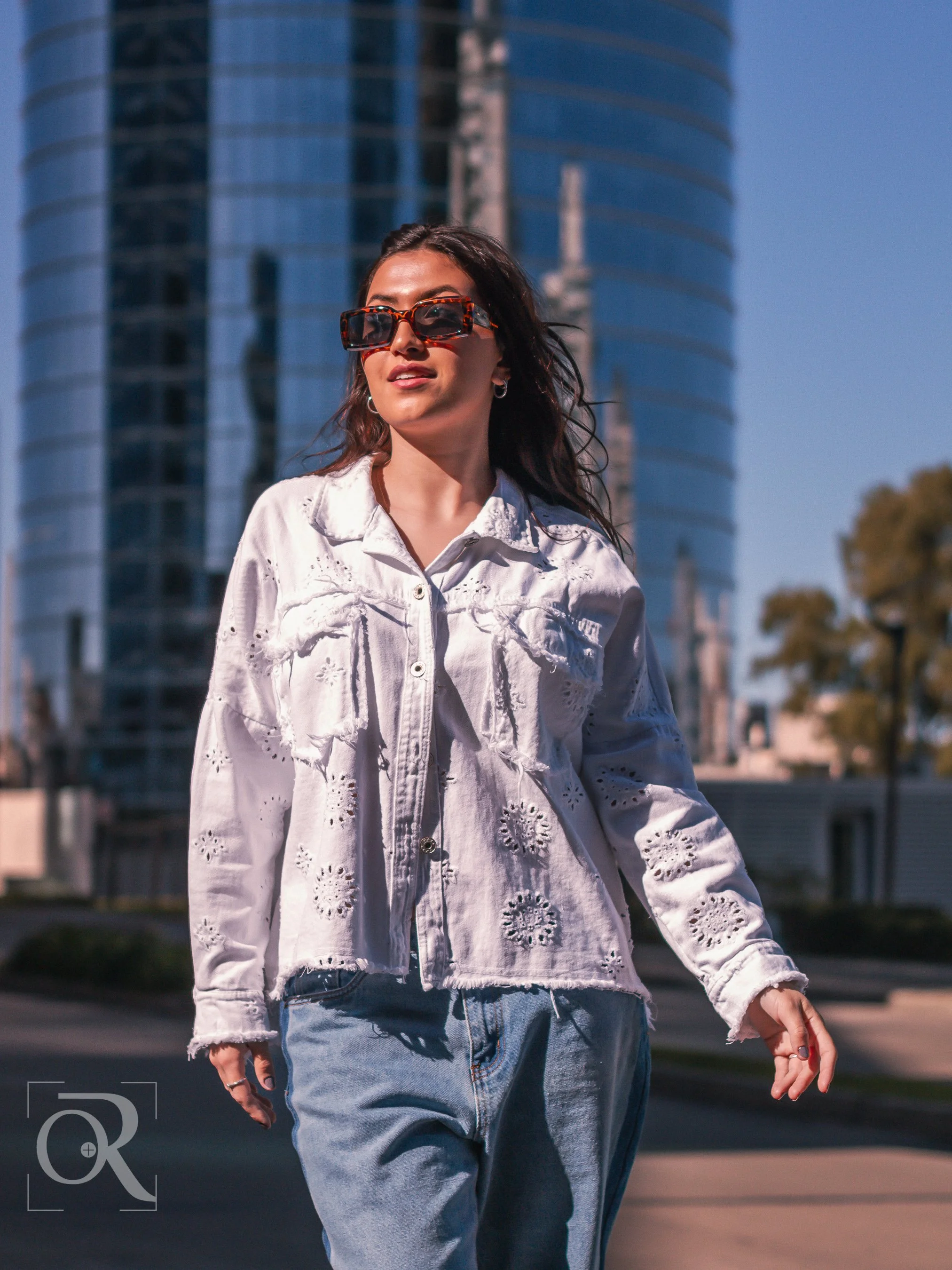 Una mujer caminando con gafas grandes de sol en un entorno urbano con edificios altos y un cielo despejado.