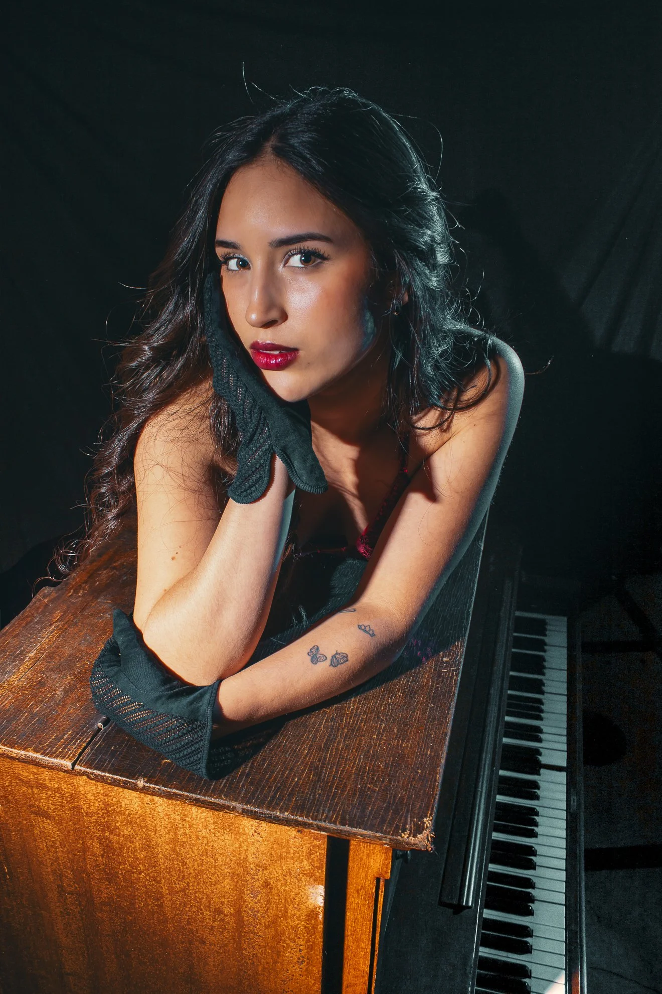 Joven mujer con cabello rizado y labios rojos, apoyada en un piano de madera, sosteniéndose una mano en la cara, con tatuajes de mariposas en el brazo, en un fondo oscuro.