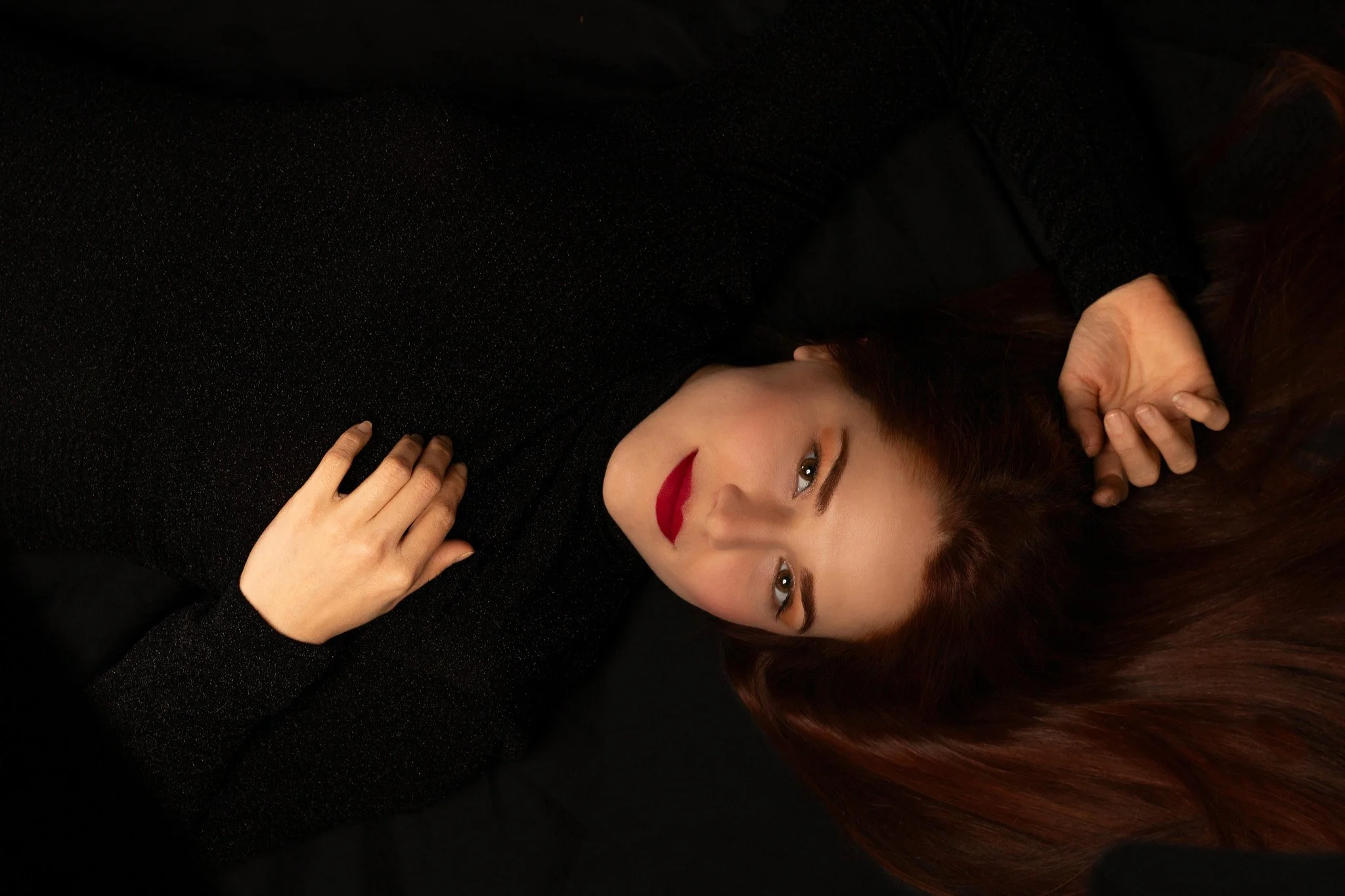Retrato de una mujer con cabello largo y rojo, vestida con ropa negra, acostada sobre una superficie negra, mirando hacía la cámara.
