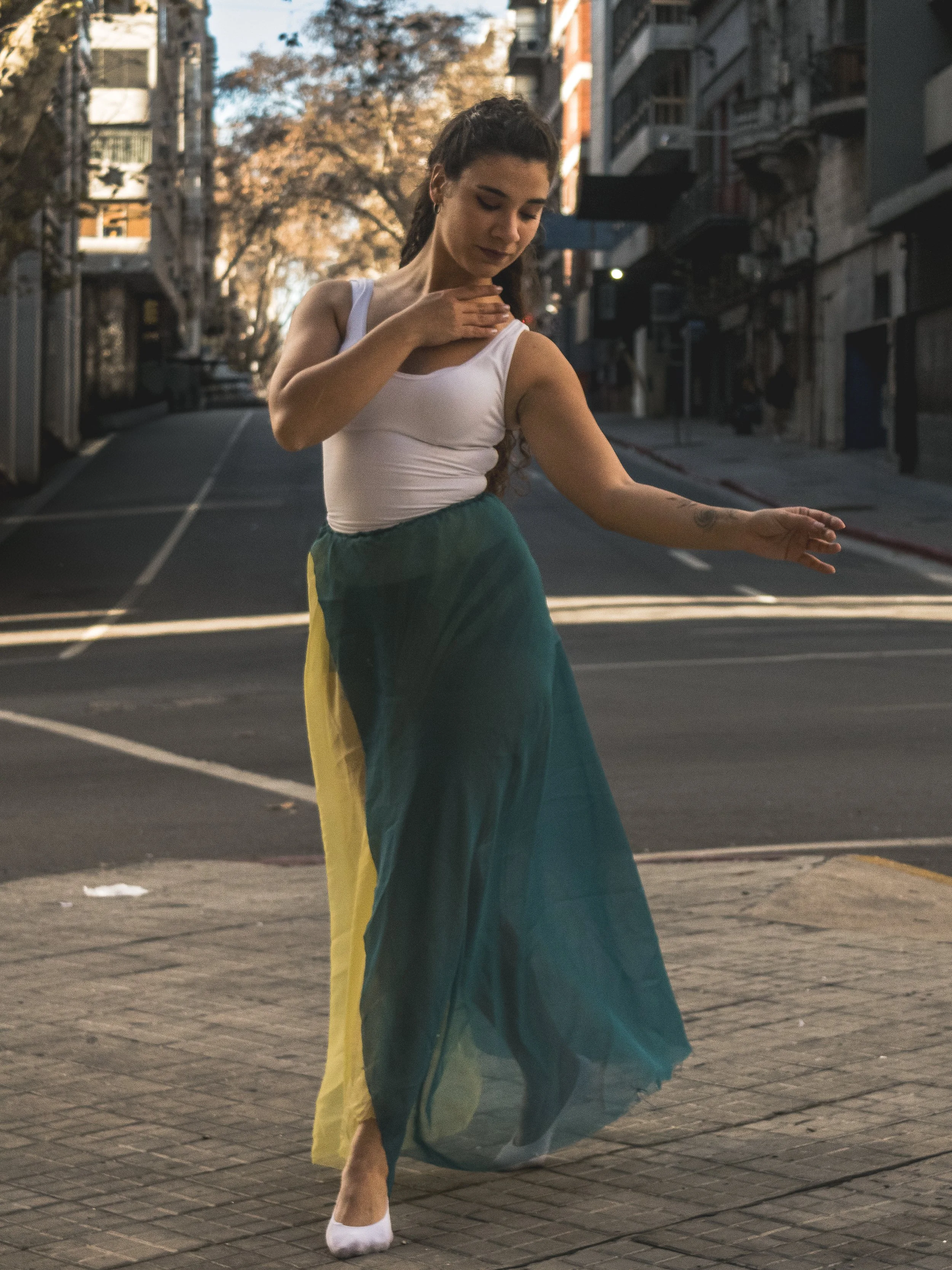 Mujer bailando en la calle con una falda larga de colores verdes, amarillos y azules, con un top blanco, en una calle urbana