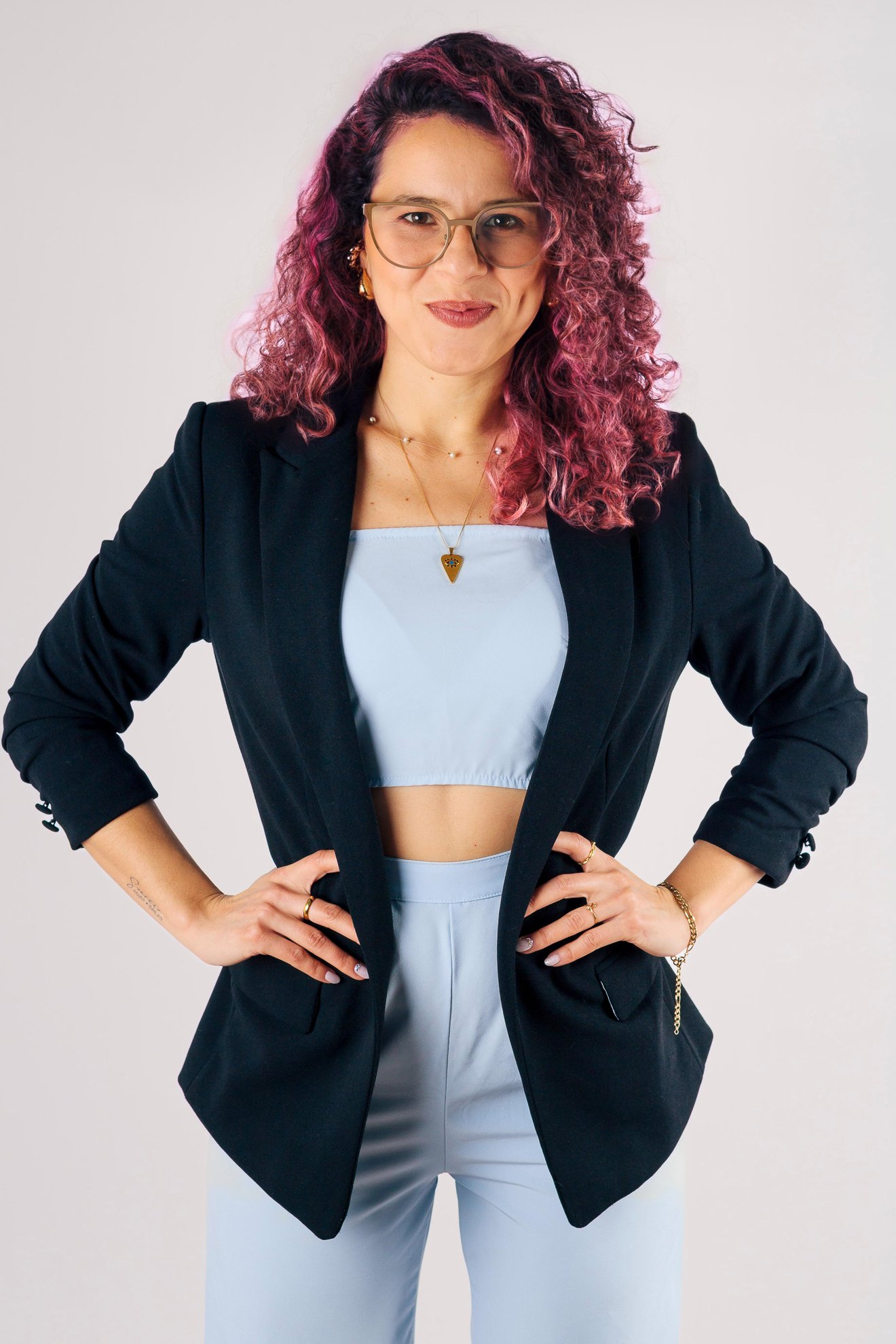 Mujer con cabello rizado y teñido de rosa, usando gafas, chaqueta negra y ropa azul claro, posa con las manos en las caderas en un fondo blanco.