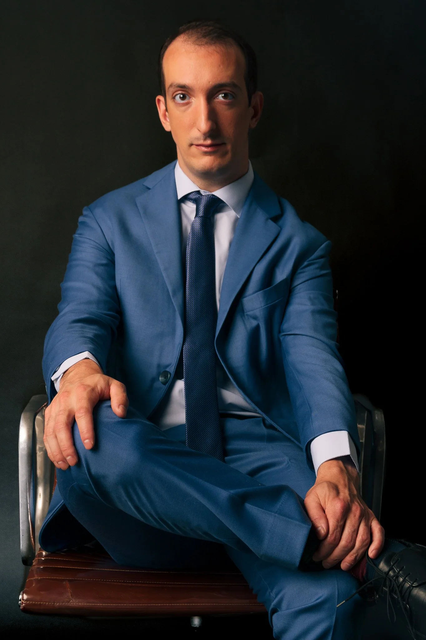 Hombre con traje azul sentado en una silla negra en un fondo oscuro.