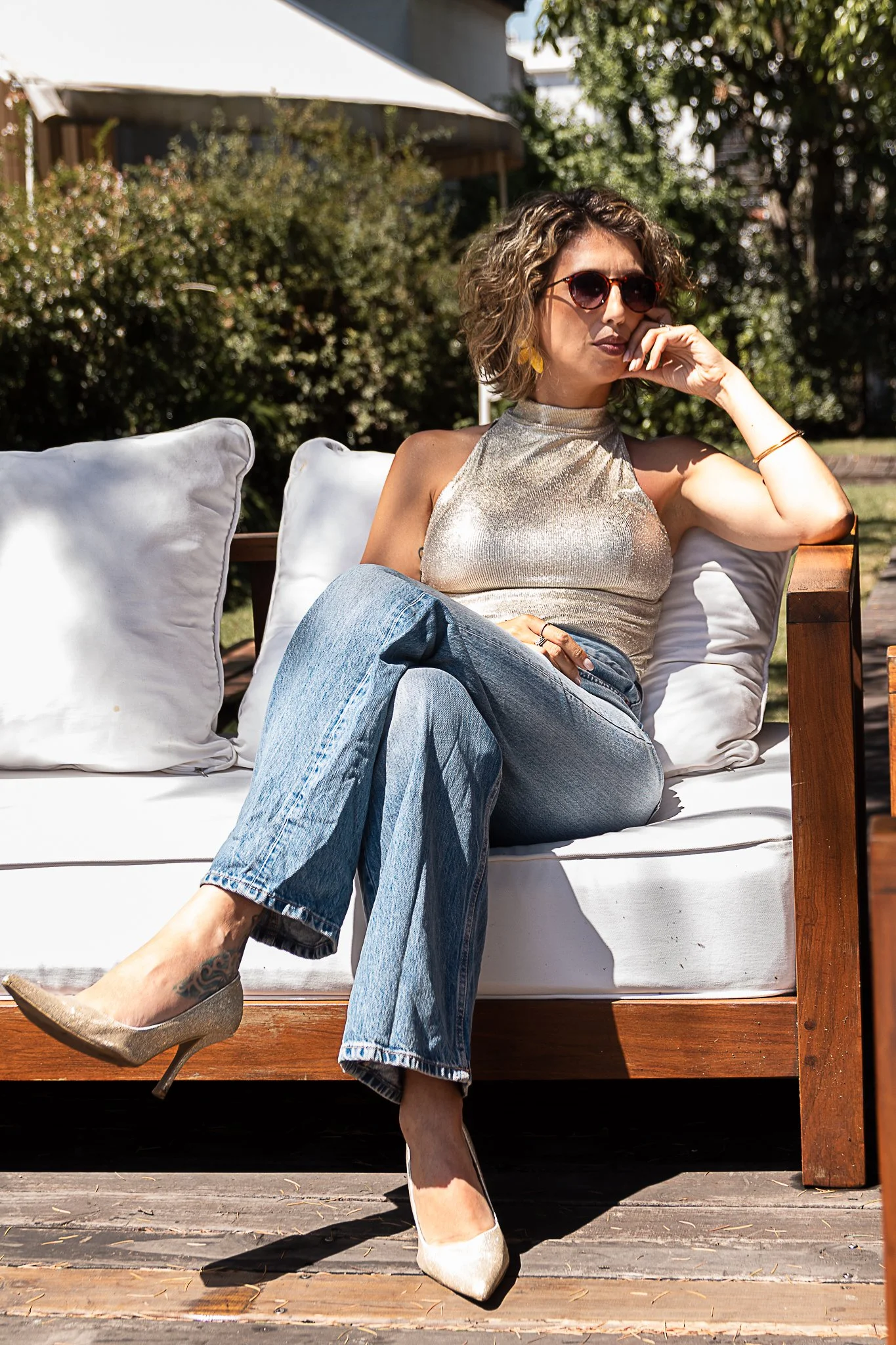 Mujer con gafas de sol y cabello rizado sentada en un sillón de madera en exteriores, vistiendo blusa dorada y jeans, usando tacones dorados.