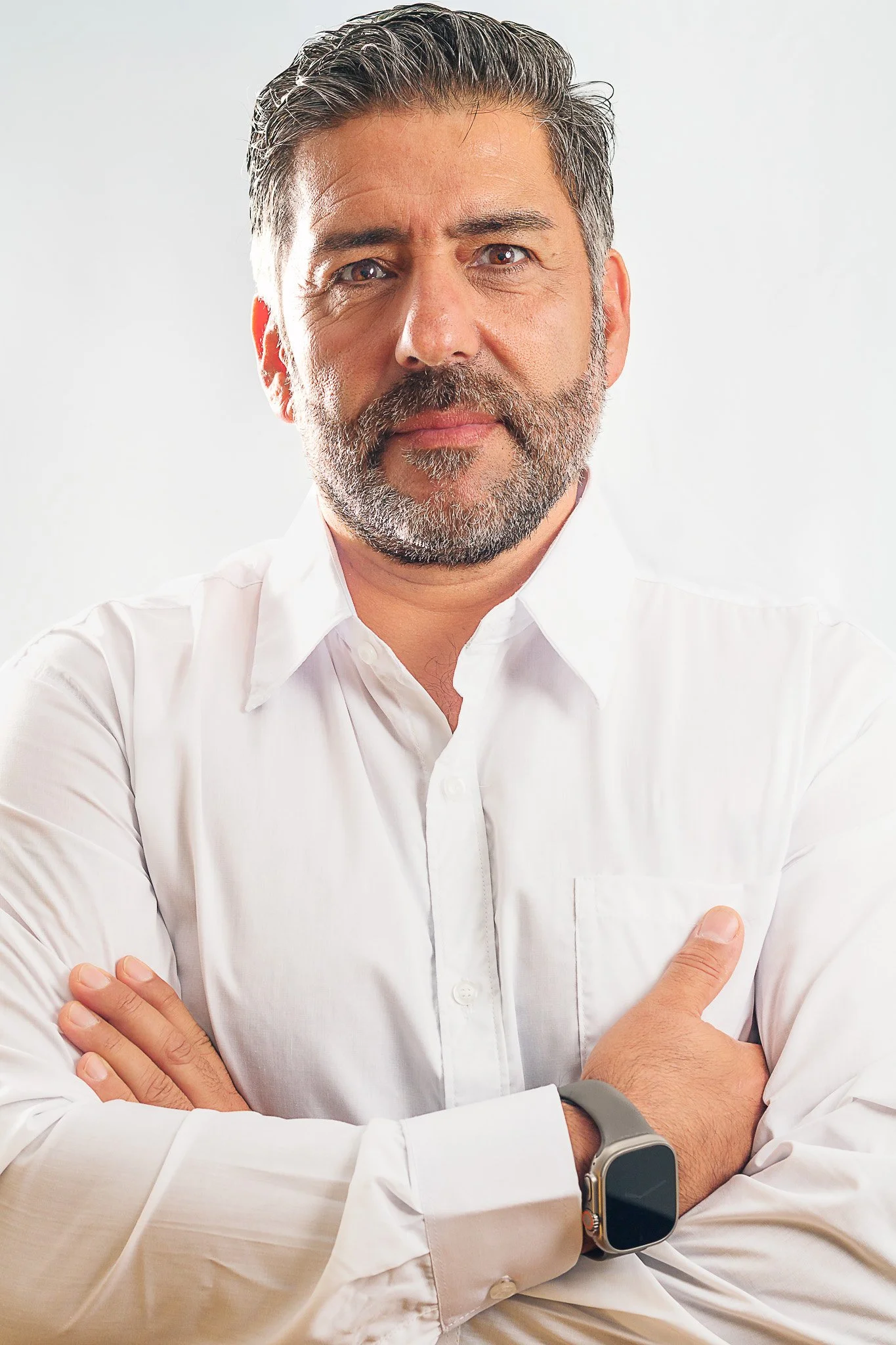 Hombre de cabello canoso y barba, usando una camisa blanca y un reloj inteligente, con los brazos cruzados, posando frente a fondo blanco.