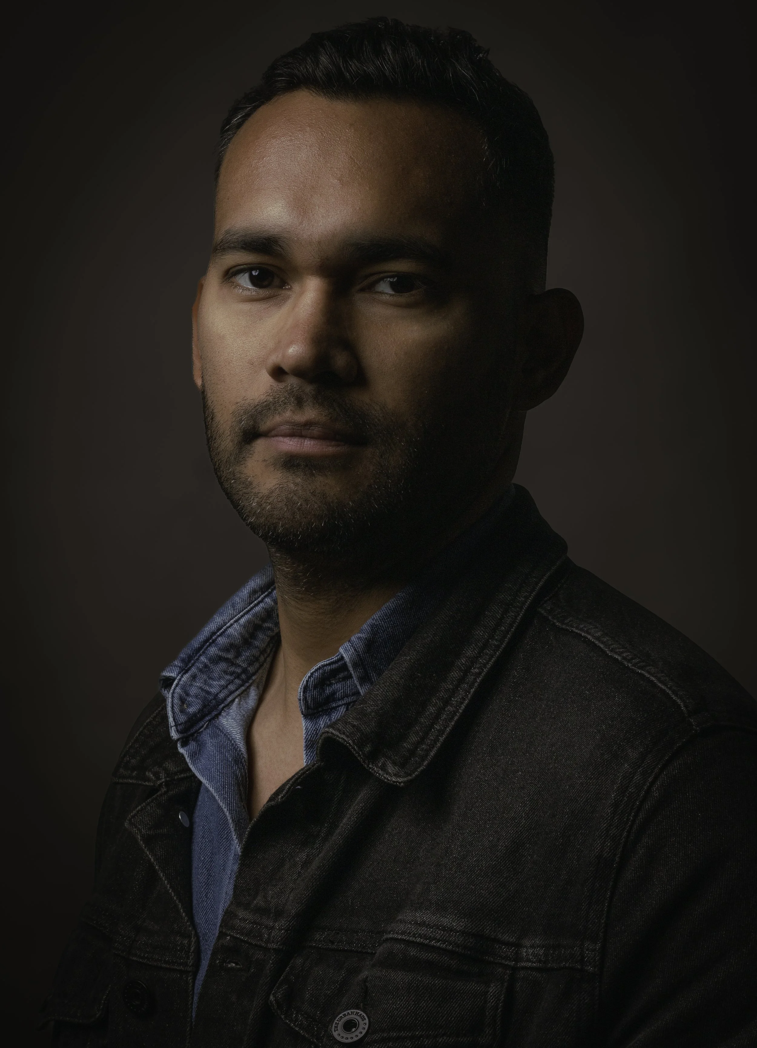 Retrato de un hombre con barba, cabello oscuro, usando chaqueta negra y camisa azul, en fondo oscuro.