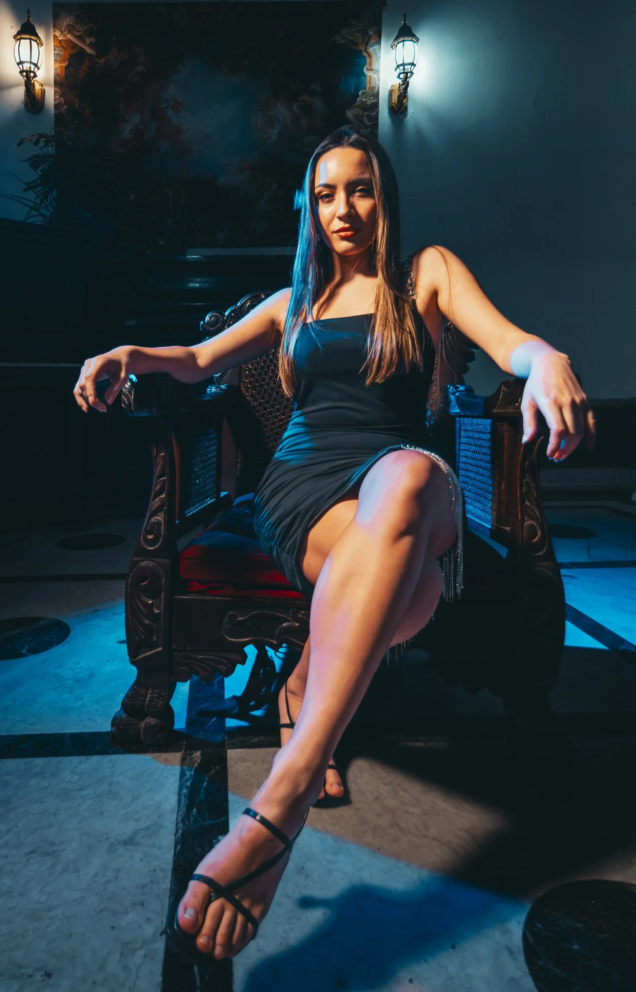 Mujer sentada en un sillón de madera, vestida con vestido negro, en un entorno con iluminación tenue y paredes con decoraciones y lámparas antiguas.