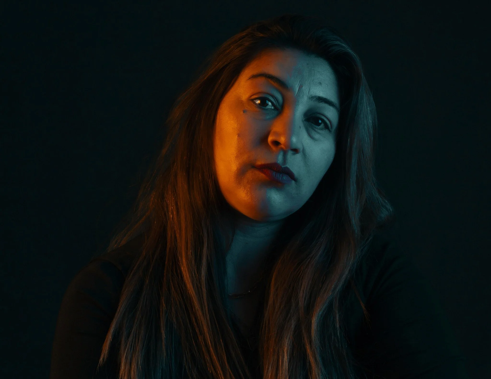 Retrato de una mujer con cabello largo y oscuro, mirada seria, fondo oscuro, iluminación de tonos cálidos y fríos en su rostro.