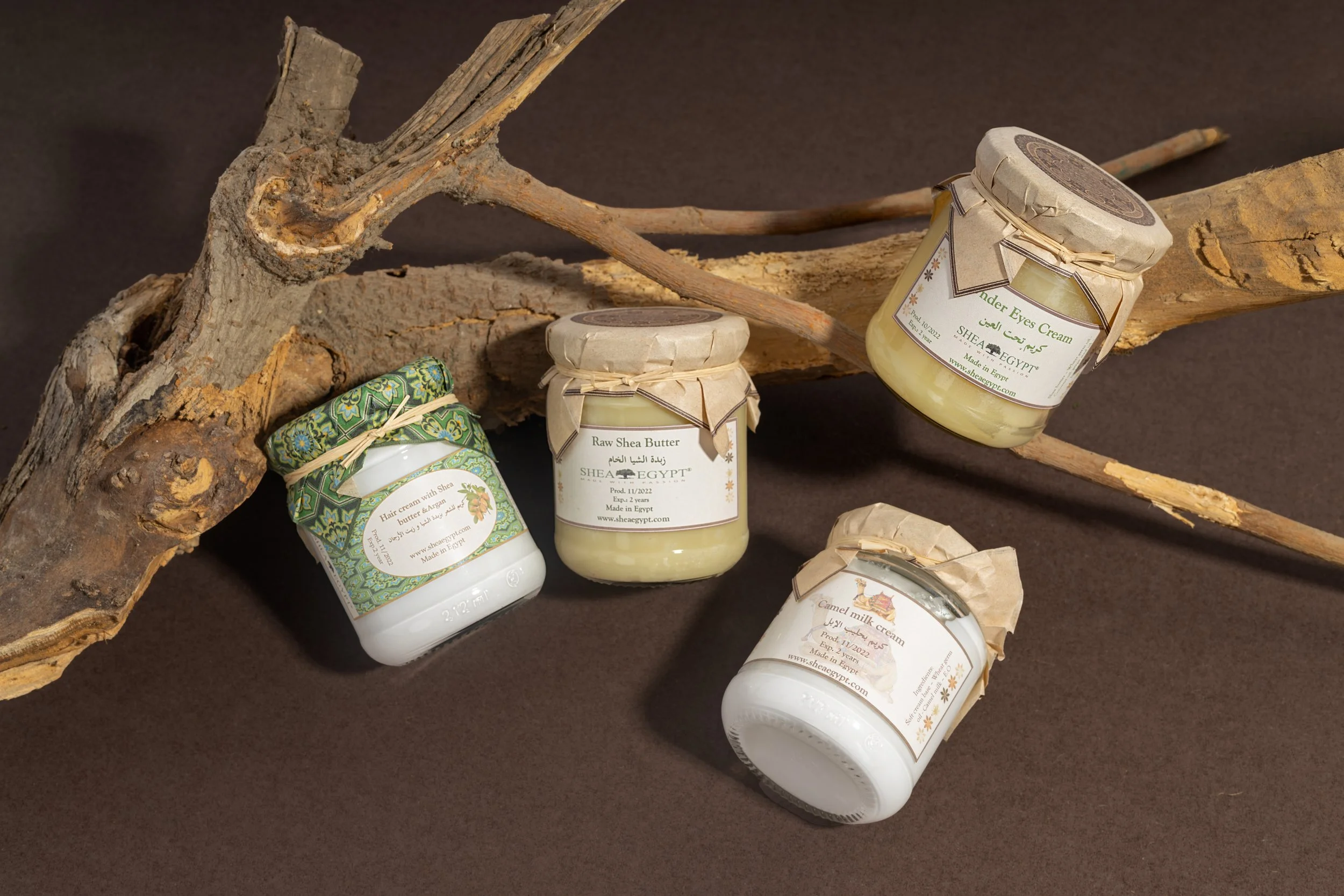 Four jars of natural skincare products, including shea butter, under eye cream, hair and camel milk cream, placed on a dark surface with a piece of wood and dry branches.