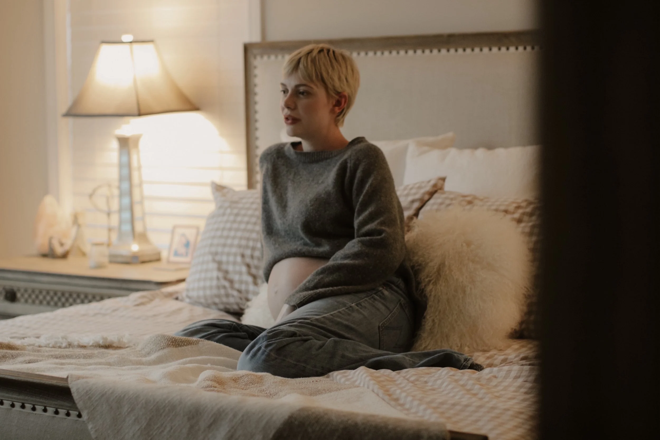 A person sitting on a bed in a cozy bedroom, wearing a gray sweater, with warm lighting and decorative pillows.