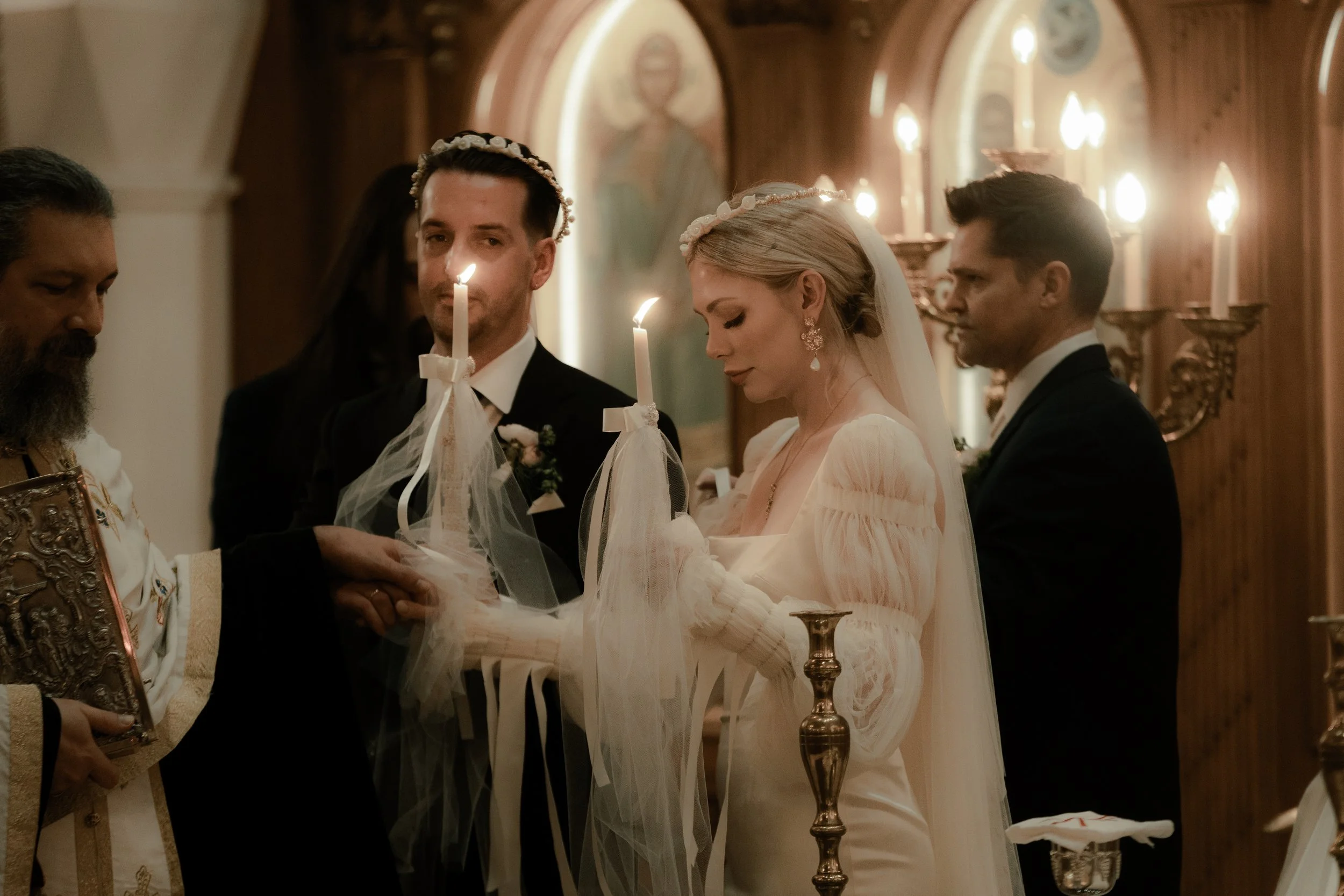 A wedding ceremony taking place inside a church, with a bride and groom holding hands and being blessed by an officiant. The bride is dressed in a white gown with wedding veil, and the groom in a black suit with a white shirt. Candles are held by the bride and groom, and there are lit candles on the wall behind them, with a religious icon or painting visible in the background.