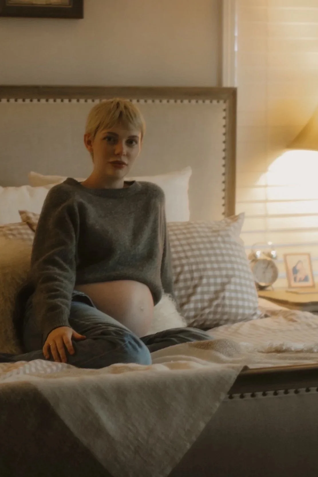 A woman with short blonde hair sitting on a bed, wearing a gray sweater and gray pants, in a cozy bedroom with soft lighting.