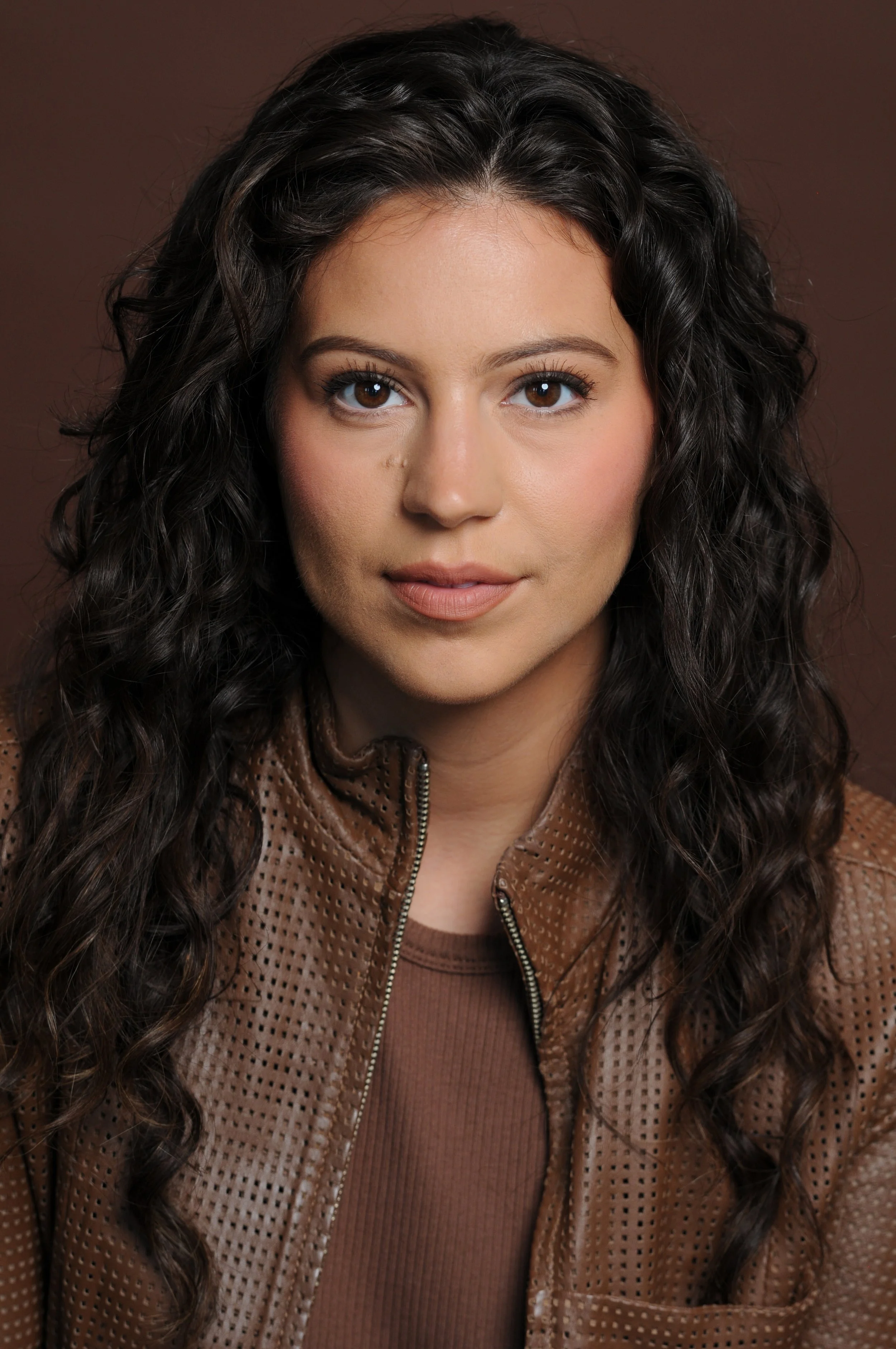 Portrait of a woman with curly dark hair, light skin, wearing a brown zip-up leather jacket and a brown shirt, against a neutral background.